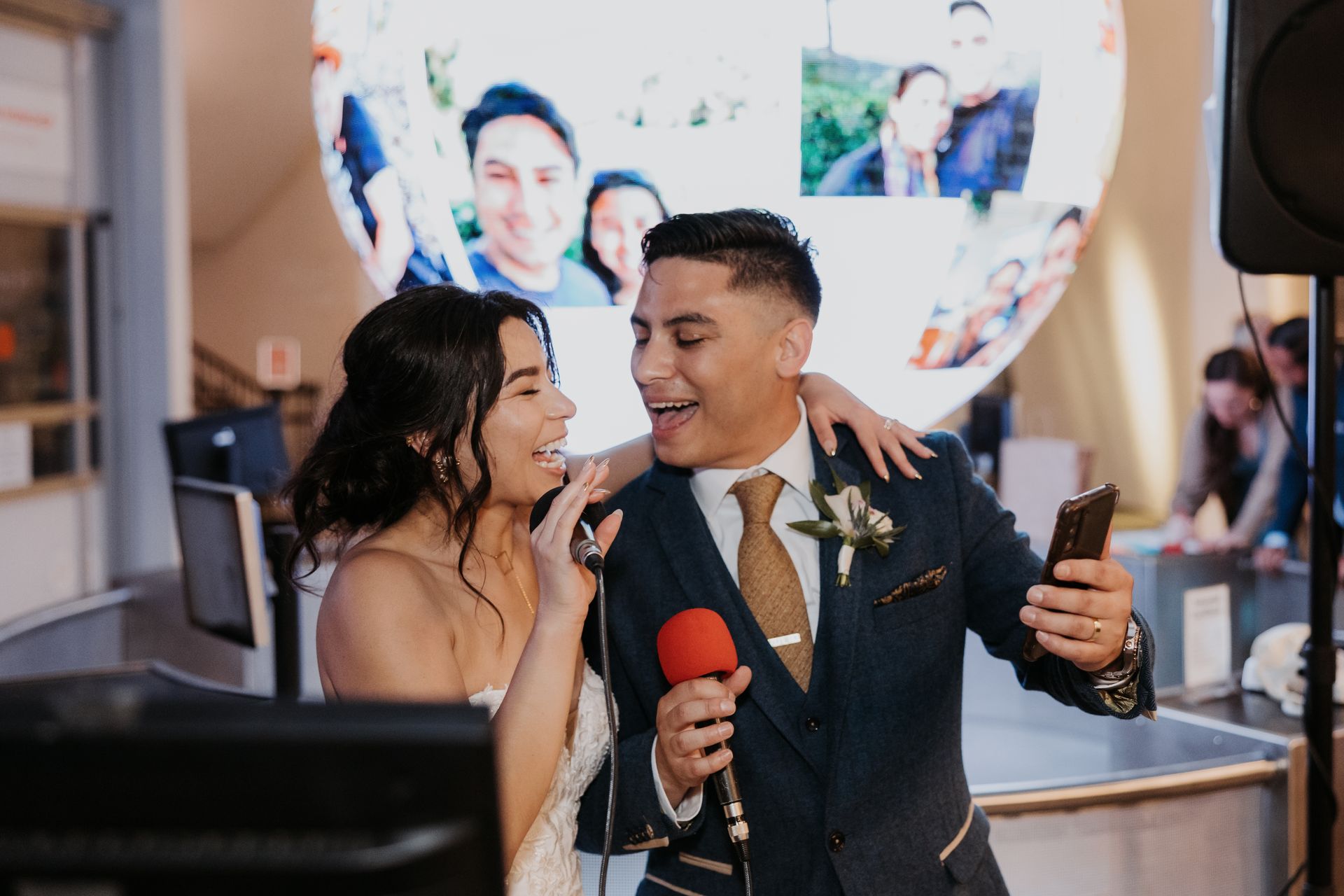 Couple in formal attire smiling at a reception, holding microphones and a drink, with a large screen behind them.