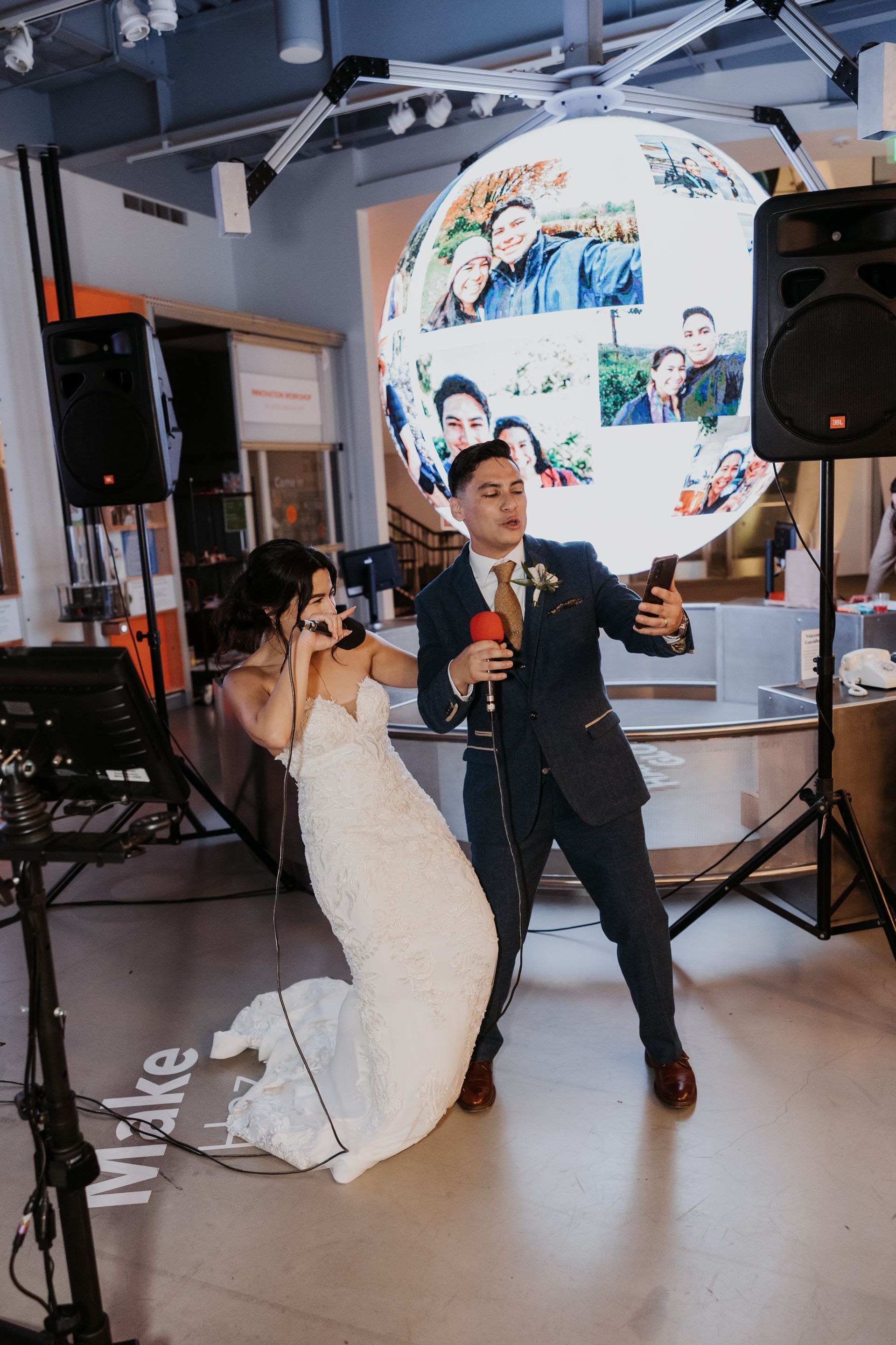 Bride and groom posing with microphones under a circular photo display at an indoor wedding reception.