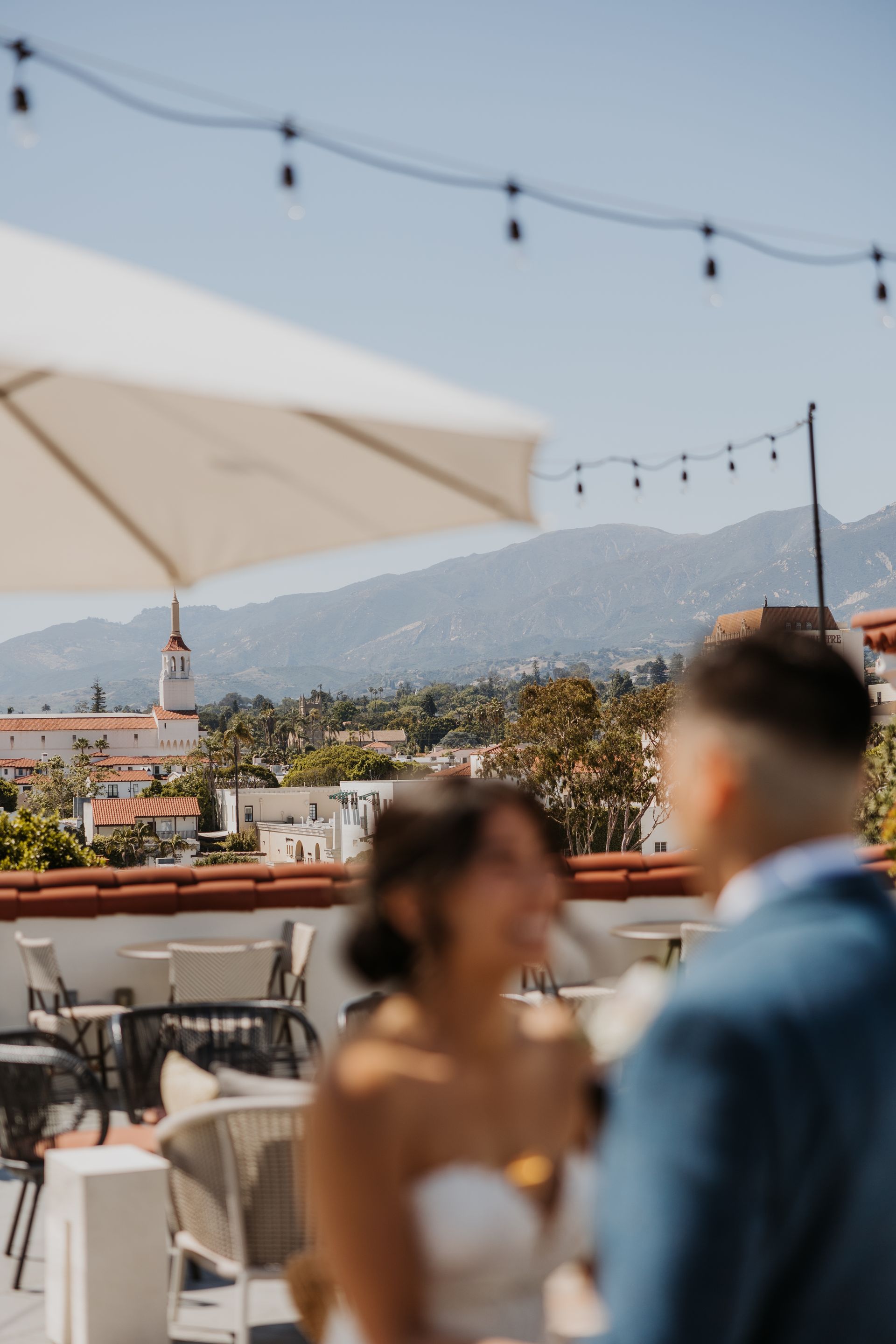 Blurred couple on a rooftop terrace with string lights, umbrella, and city and mountain views