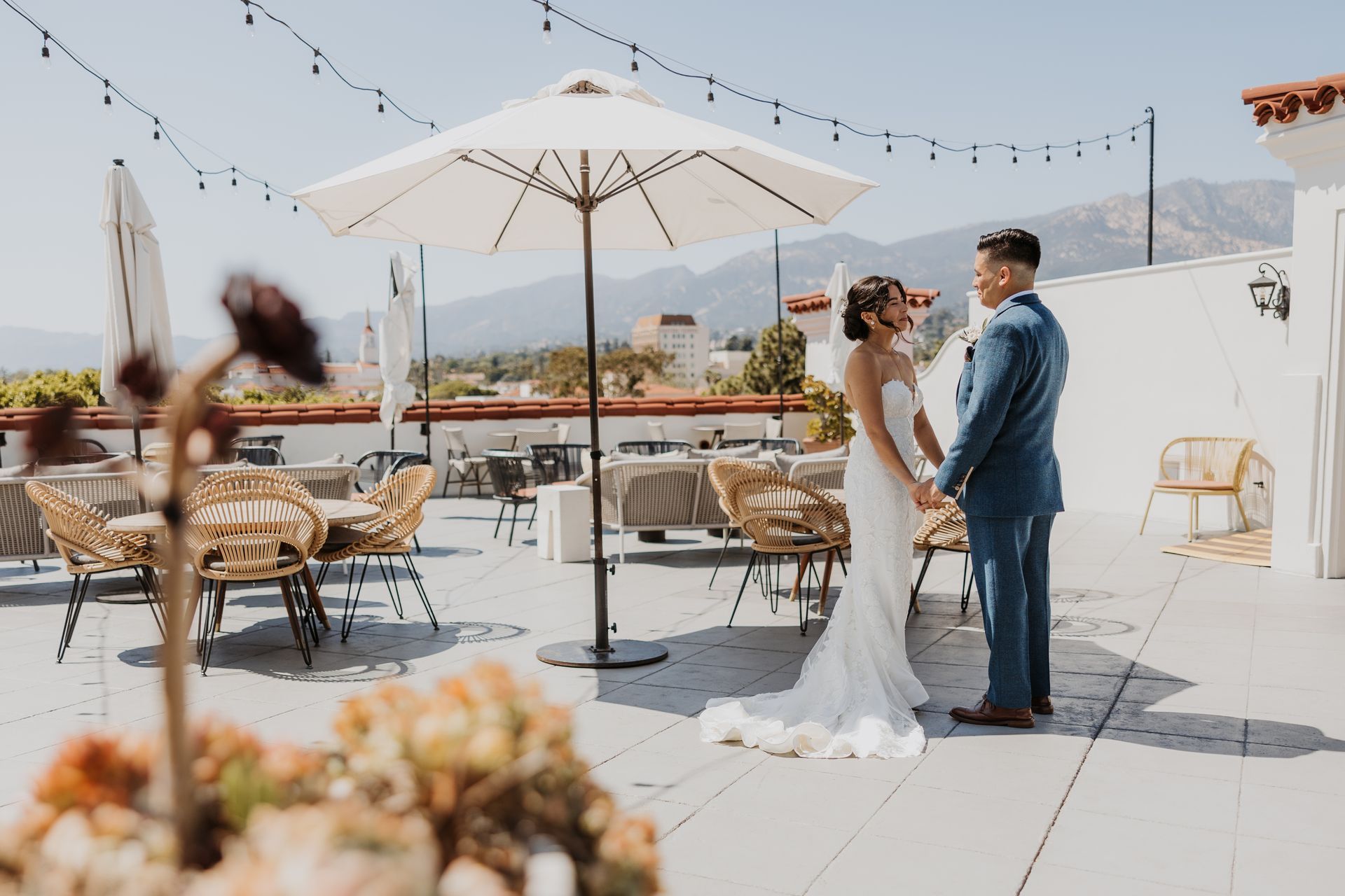 Bride and groom standing on a rooftop patio with umbrellas and string lights, holding hands in wedding attire
