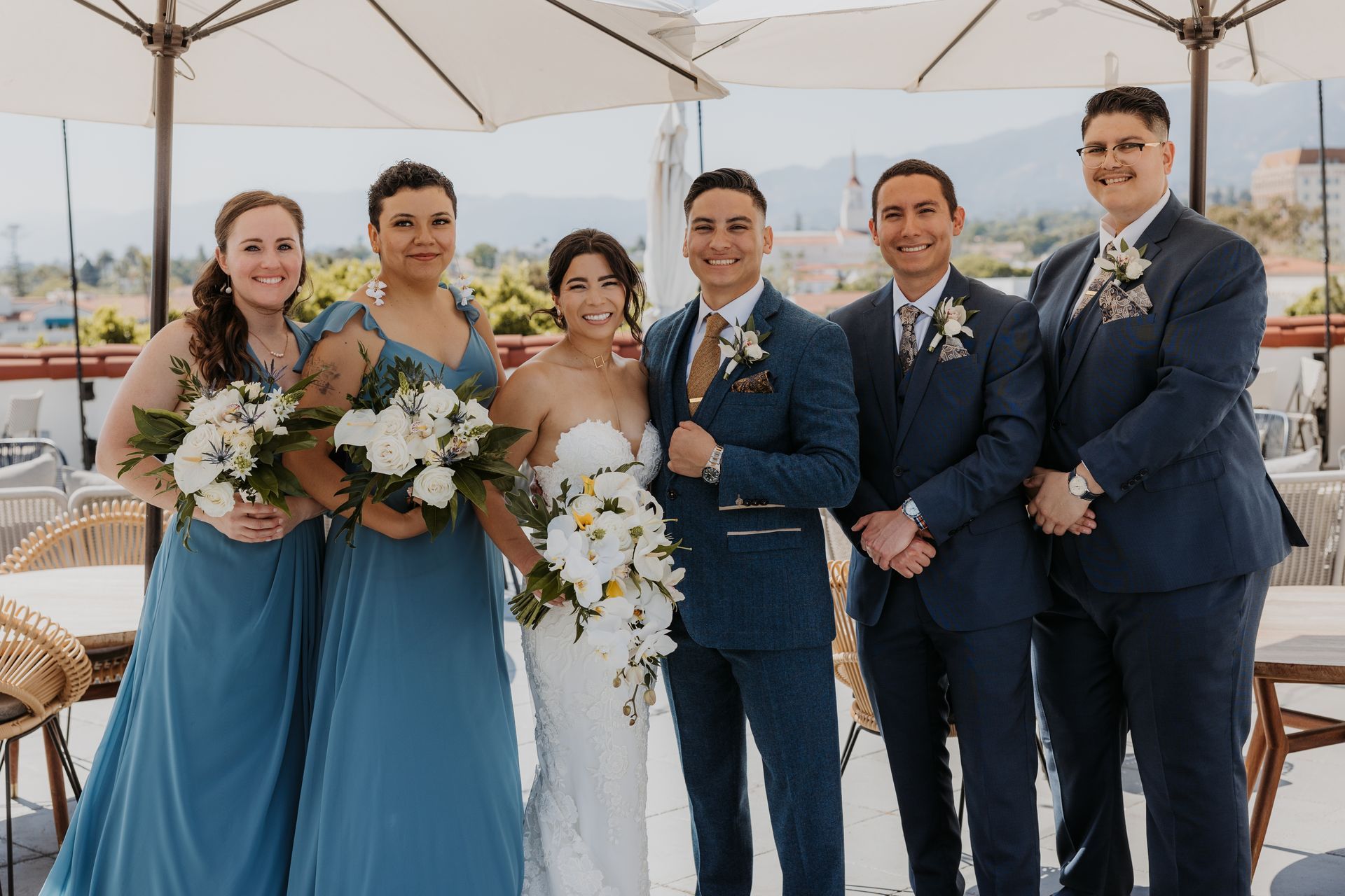 Wedding party posing outdoors under umbrellas, with two women in blue dresses, a bride, and three men in navy suits.