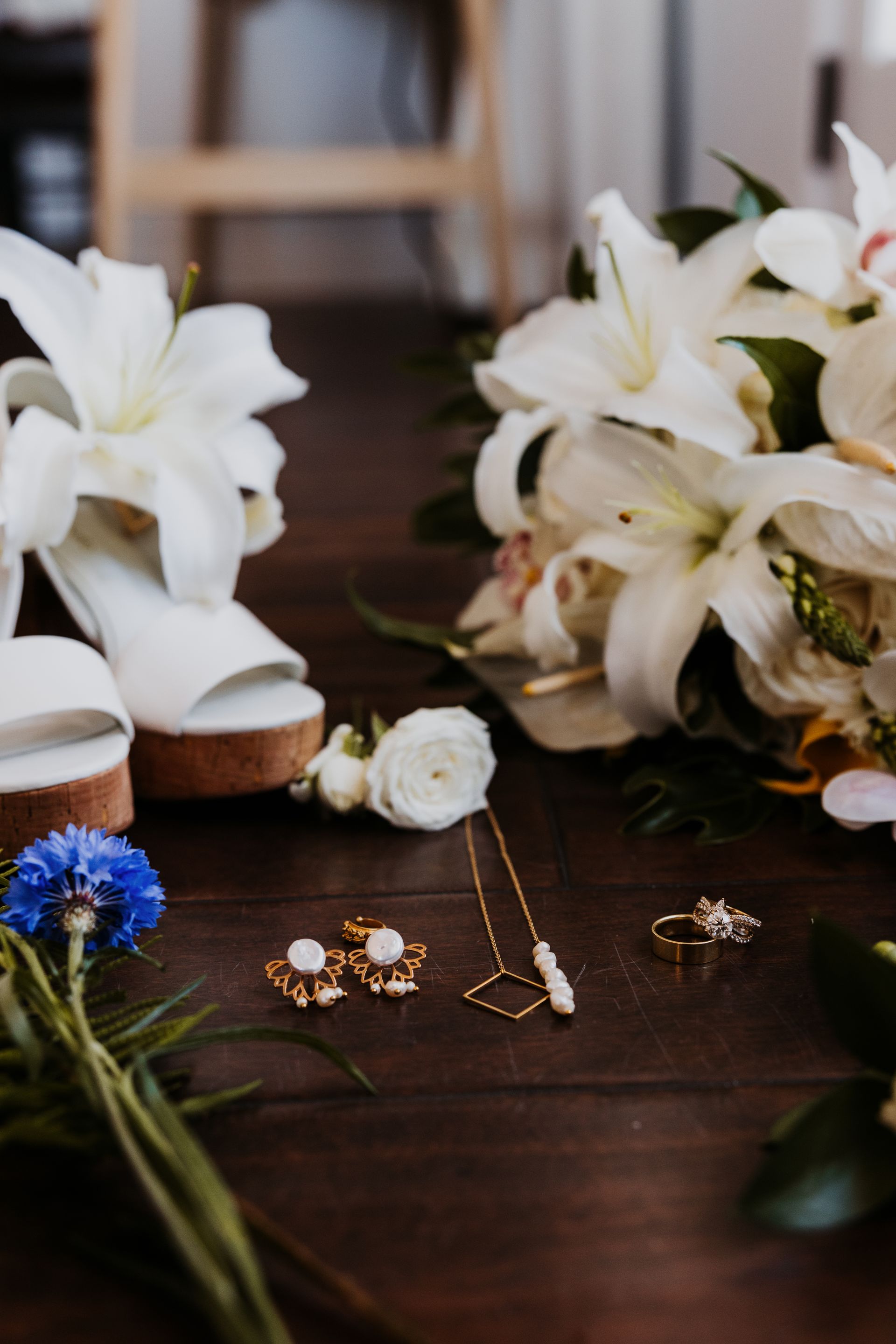 Wedding jewelry and white lilies arranged on a dark wooden table with white shoes in the background