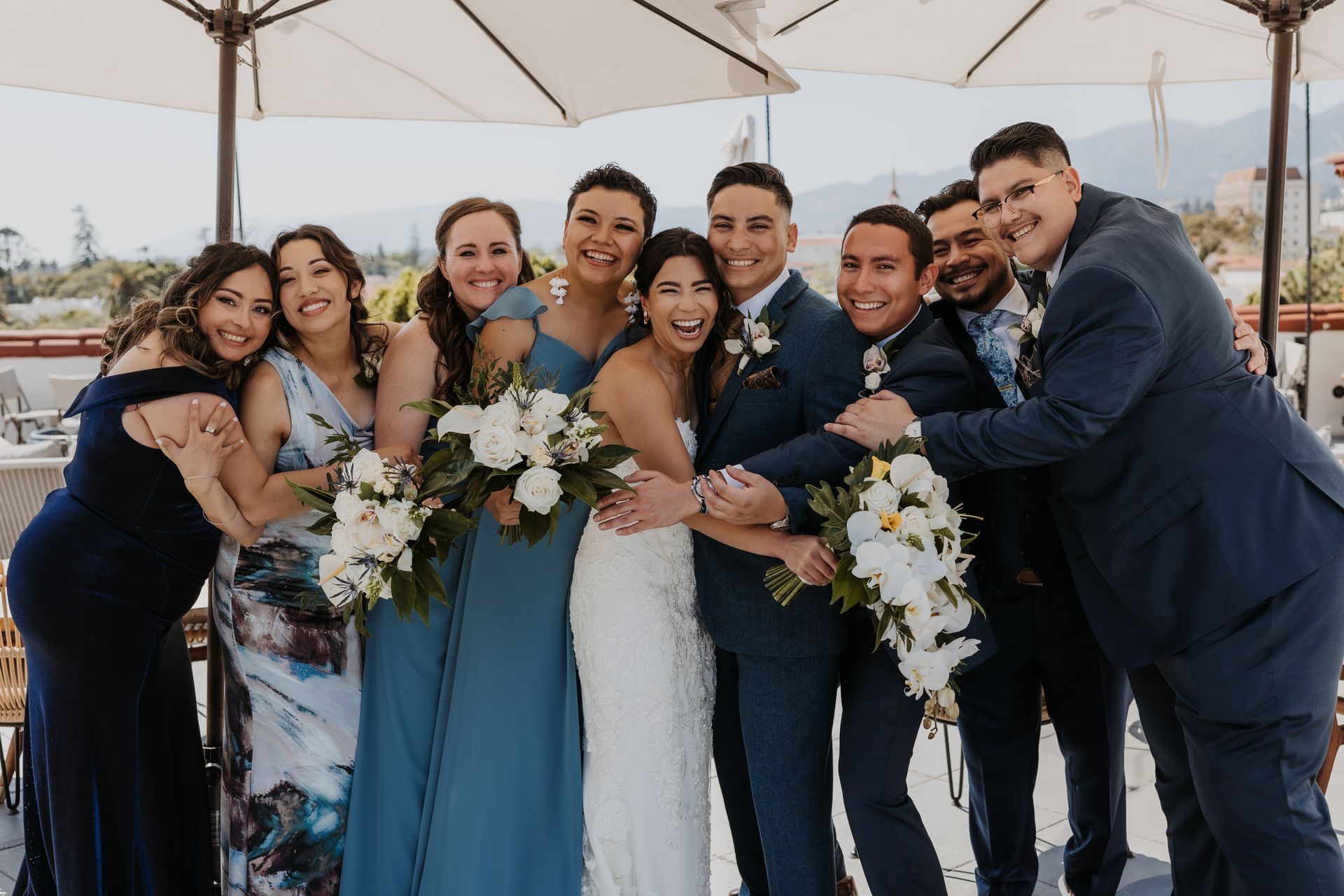 Wedding party posing together outdoors under umbrellas, with the bride in a white dress and others in formal attire.