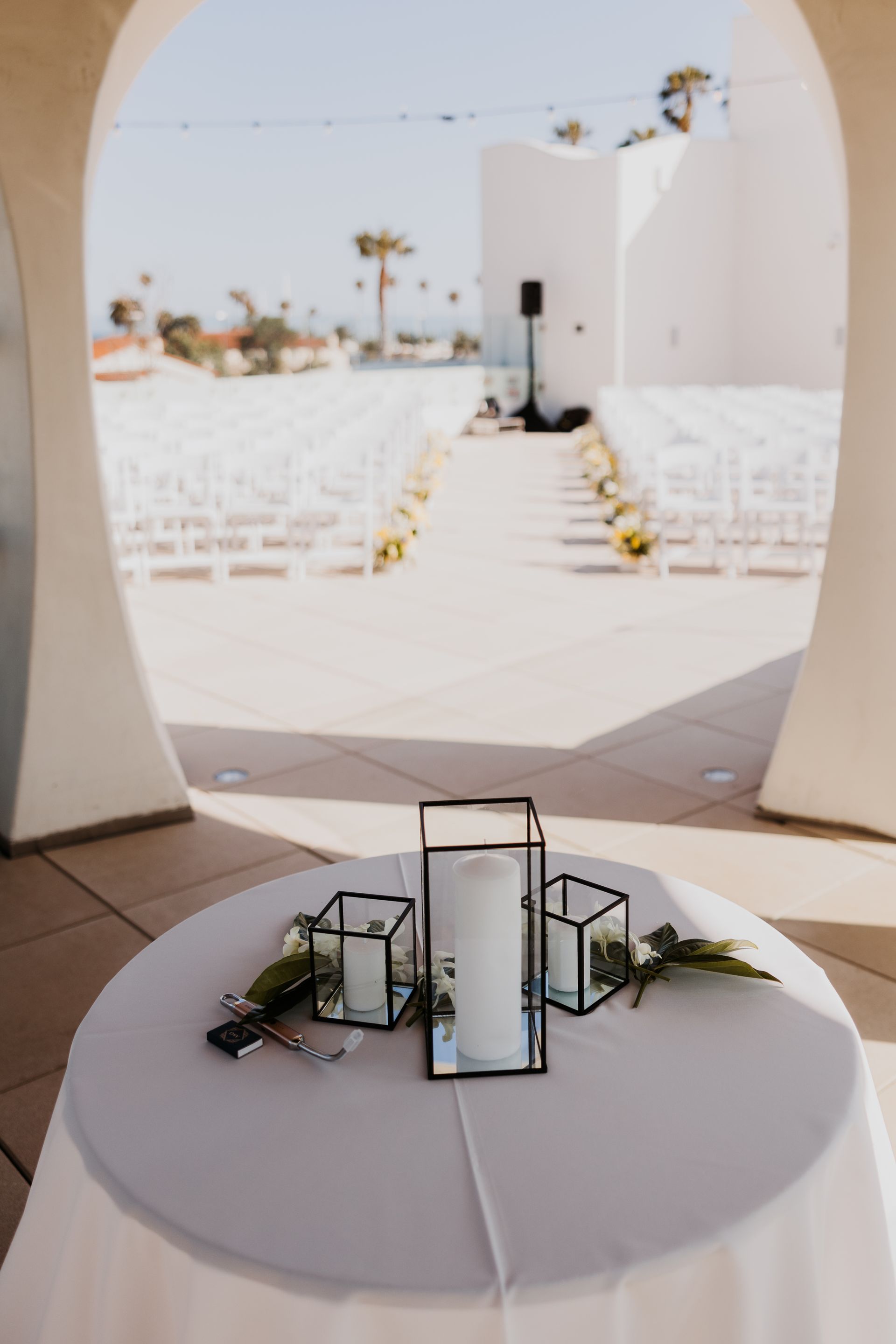 White-clothed wedding aisle leading to a ceremony arch, with a round table and lantern centerpiece in the foreground.