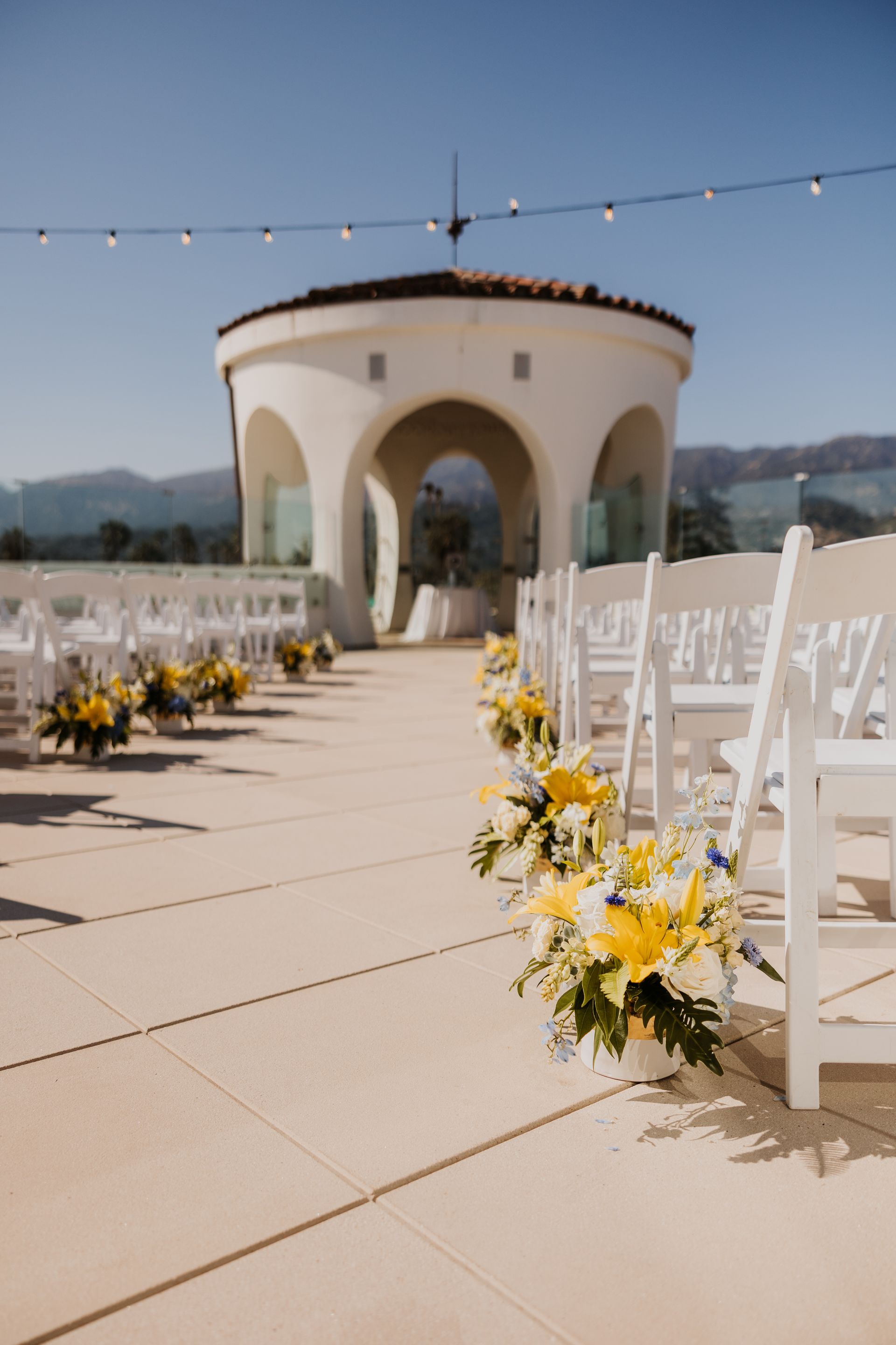 Outdoor wedding aisle with white chairs, yellow flowers, and a small archway pavilion under string lights