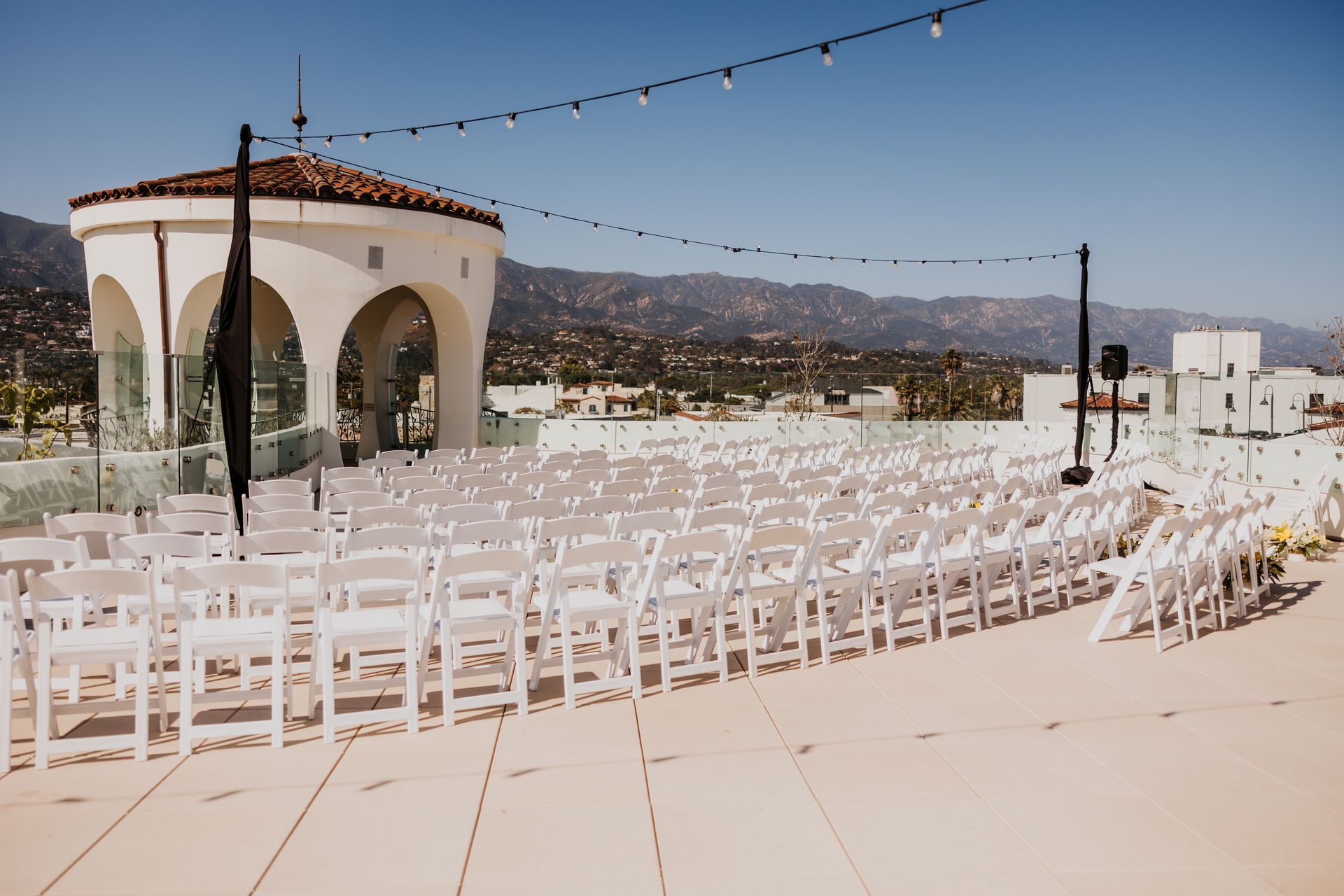 Outdoor wedding setup with rows of white chairs on a sunny terrace beside a small pavilion and mountain view