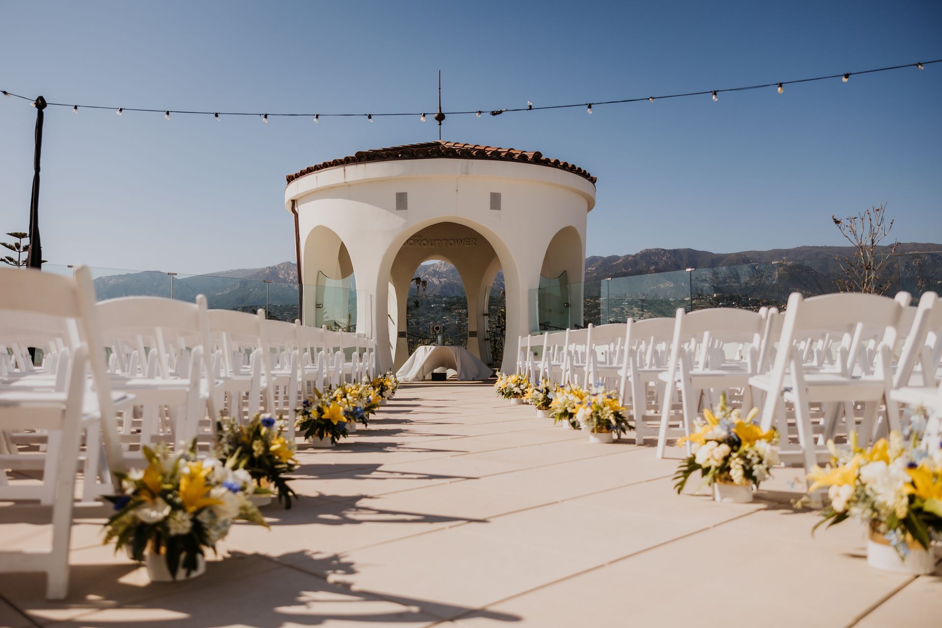 Outdoor wedding aisle with white chairs and yellow-white flower arrangements leading to a pavilion under a blue sky