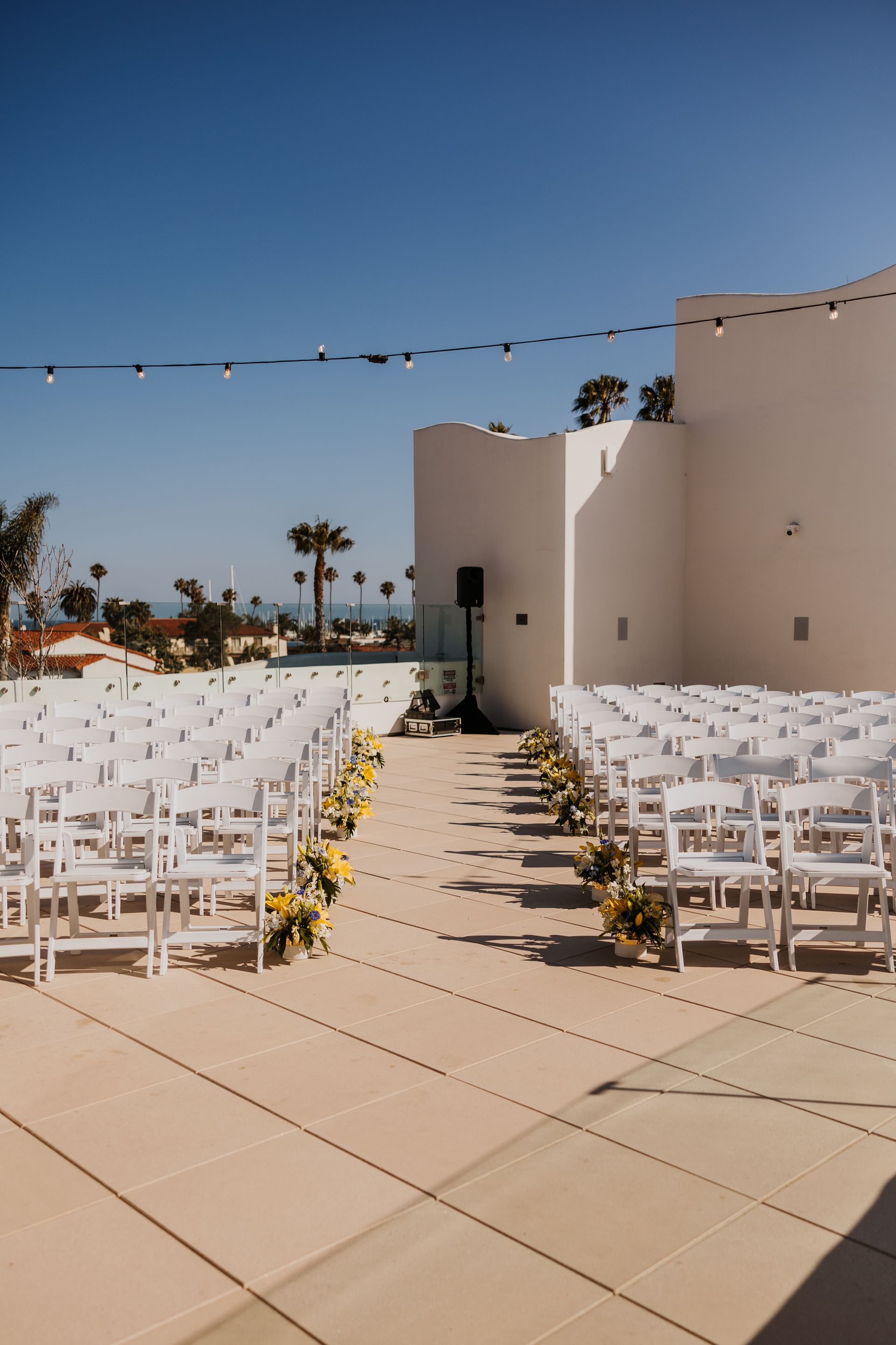 Outdoor wedding ceremony setup with rows of white chairs, flower-lined aisle, and white stucco walls under a blue sky