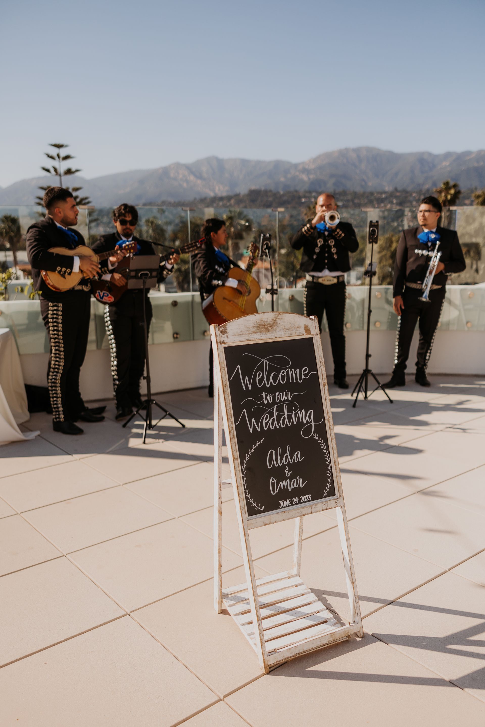 Musicians playing outdoors beside a chalkboard sign on a sunny patio, with mountains in the background