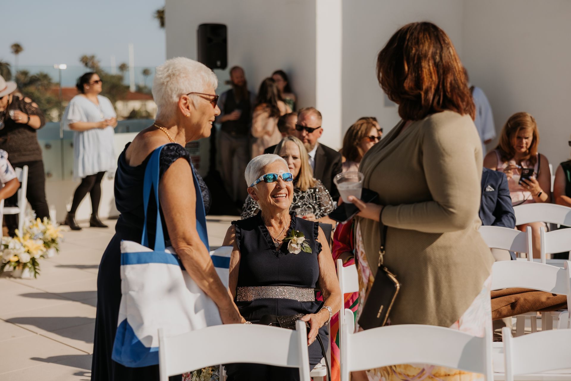 Guests chatting at an outdoor ceremony, with seated attendees and white chairs in the foreground