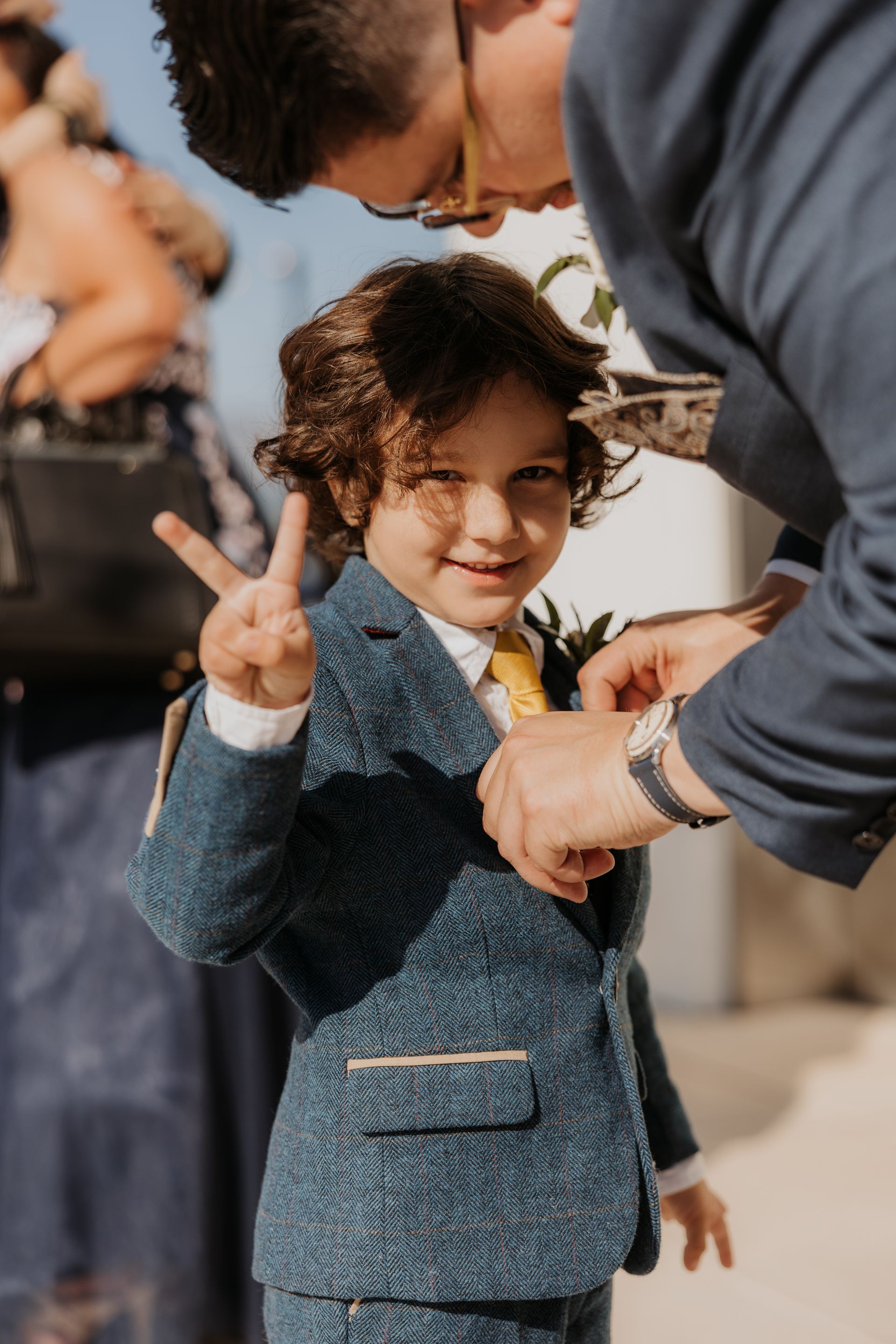 Child in a blue suit smiling and holding up a peace sign while adults adjust their clothes outdoors