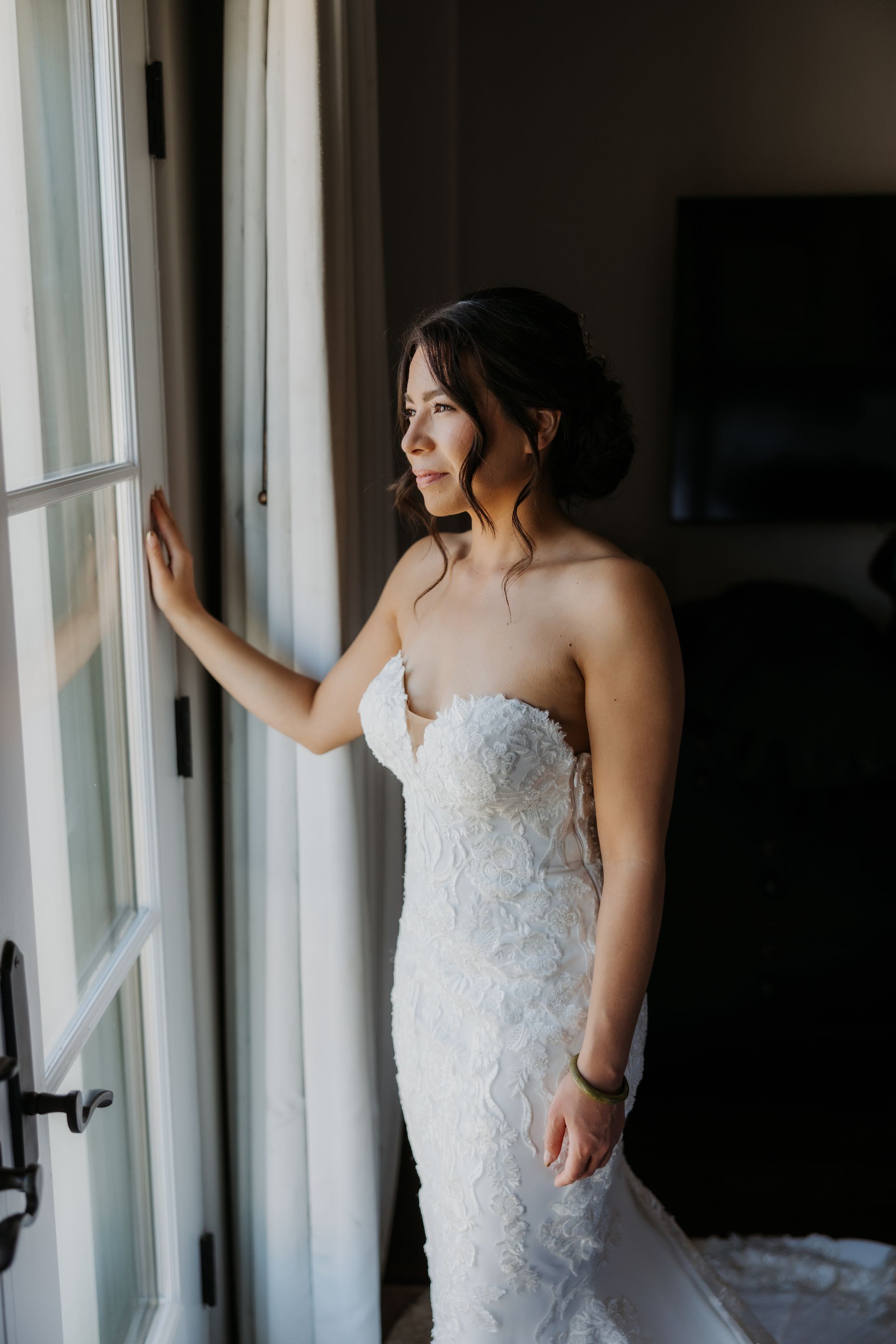 Bride in a strapless white gown standing by a window, looking outside in soft light