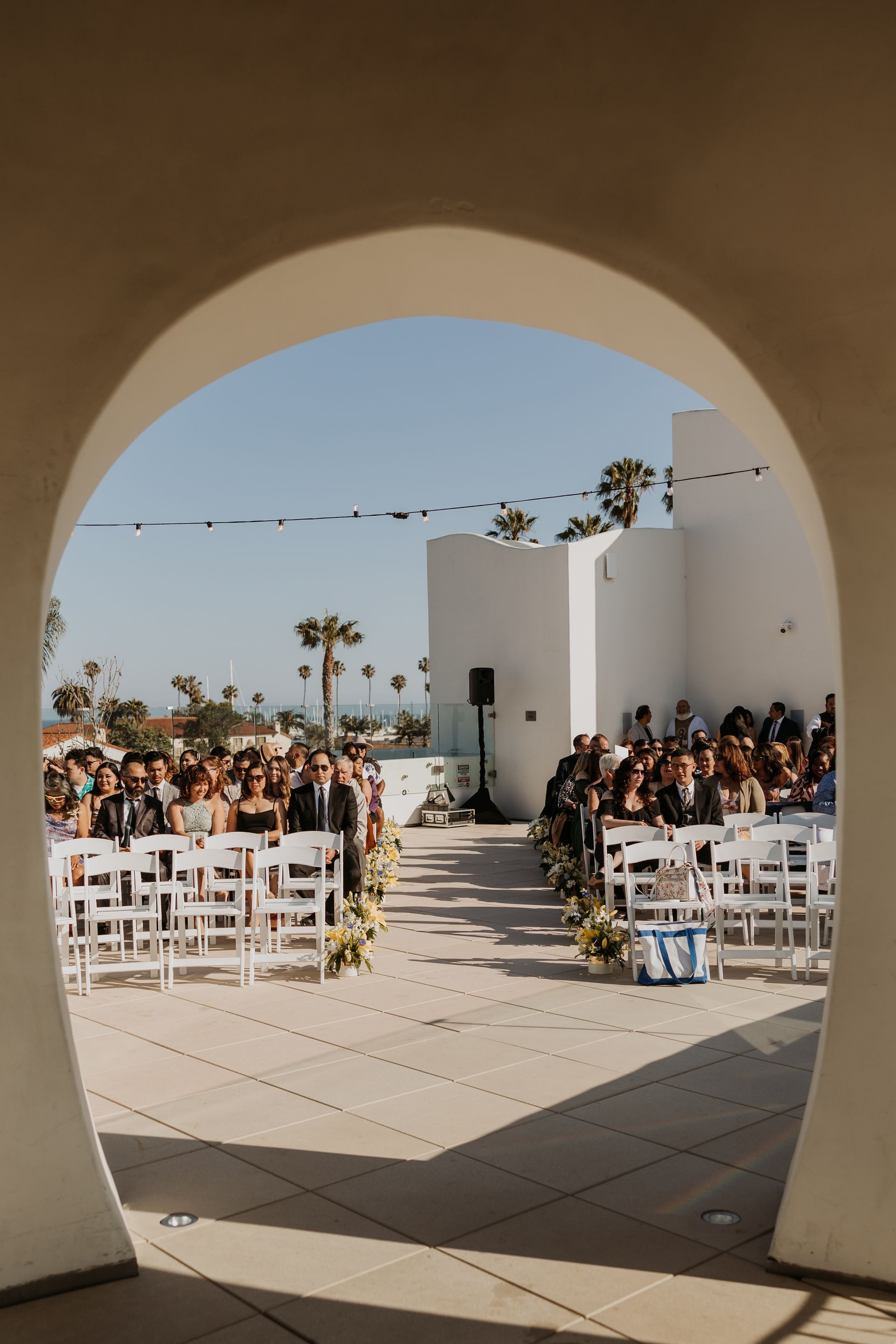 Outdoor wedding ceremony viewed through a white arch, with rows of white chairs and guests seated ahead.