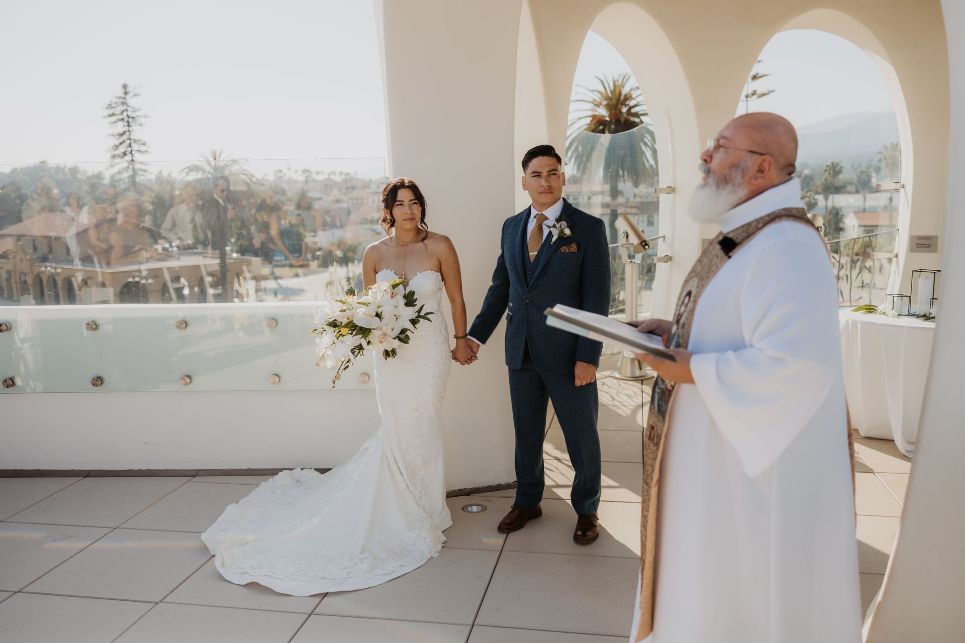Bride and groom stand with an officiant during an outdoor wedding ceremony on a terrace.