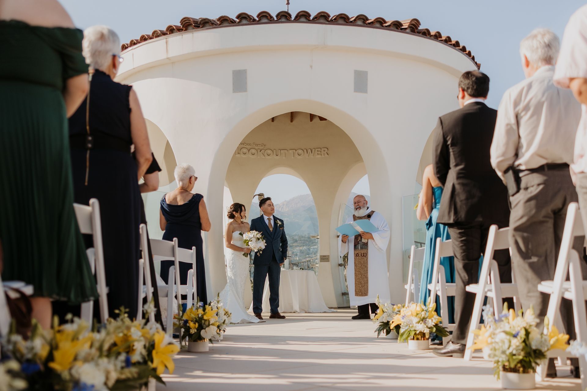 Wedding ceremony under white archway with guests seated in an outdoor aisle lined with flowers.