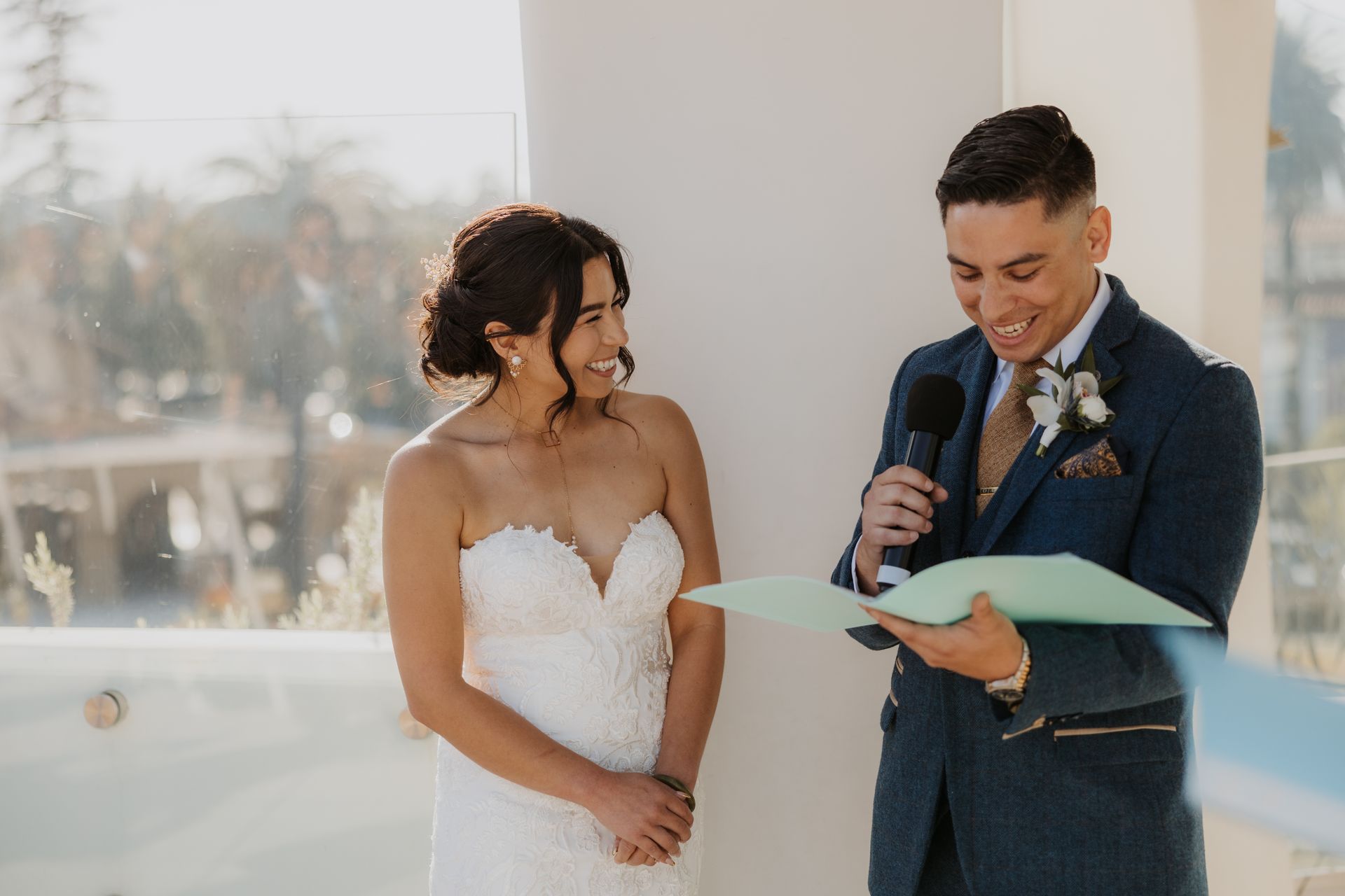 Bride smiles as groom reads vows from a card during a wedding ceremony outdoors.
