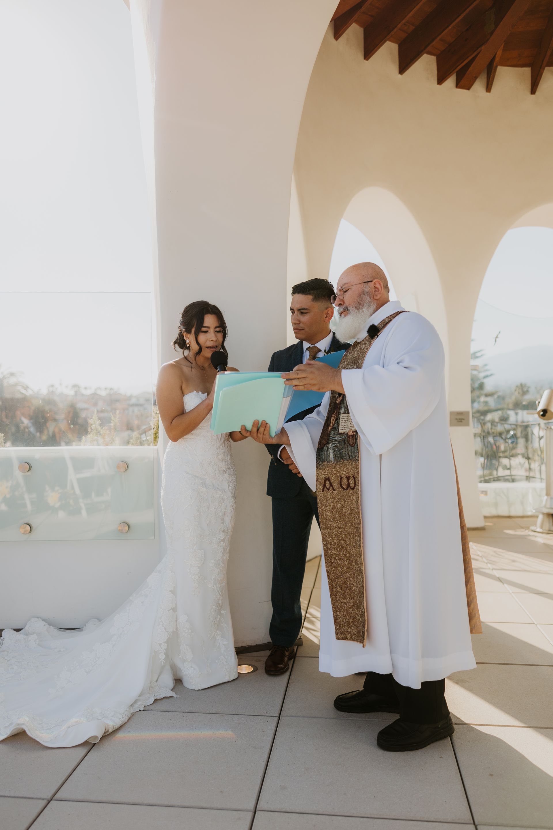 Bride, groom, and officiant reading vows on a sunlit balcony during a wedding ceremony
