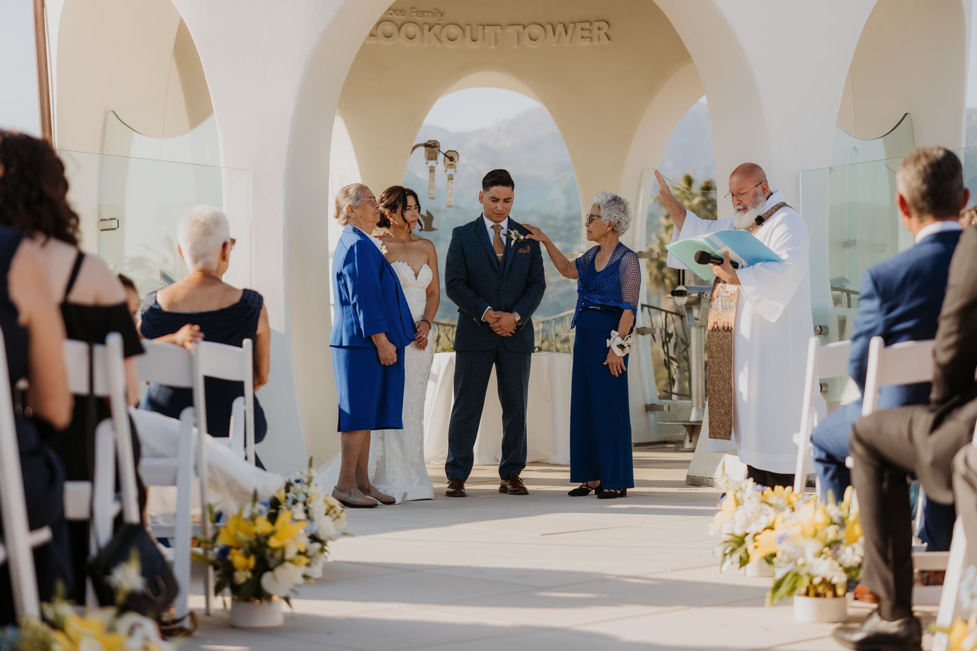 Wedding ceremony in a white arched courtyard, with guests seated and the couple standing at the altar