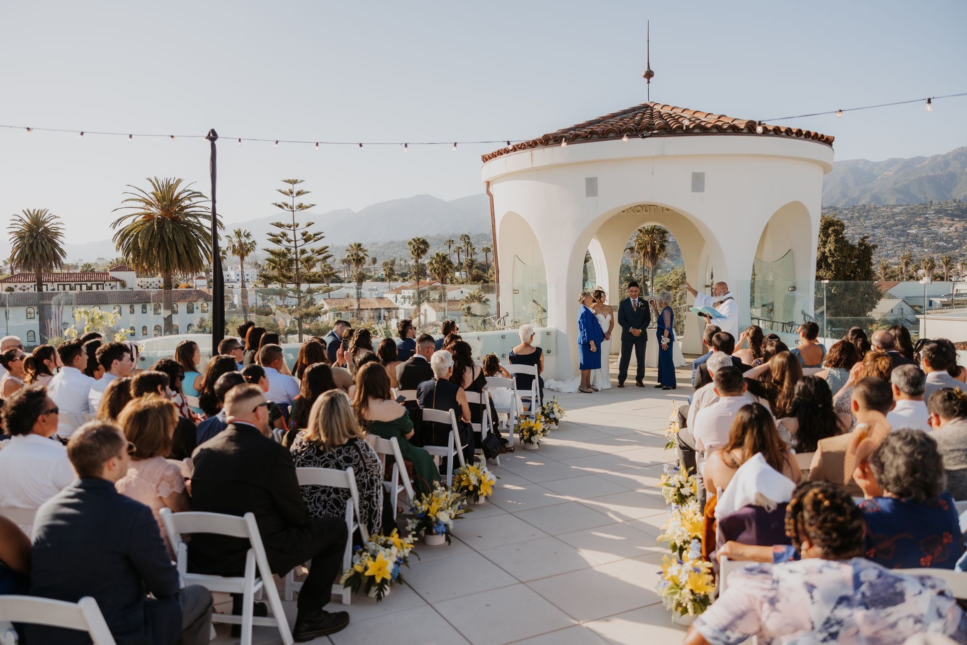 Outdoor wedding ceremony with seated guests facing a white gazebo by the water at sunset