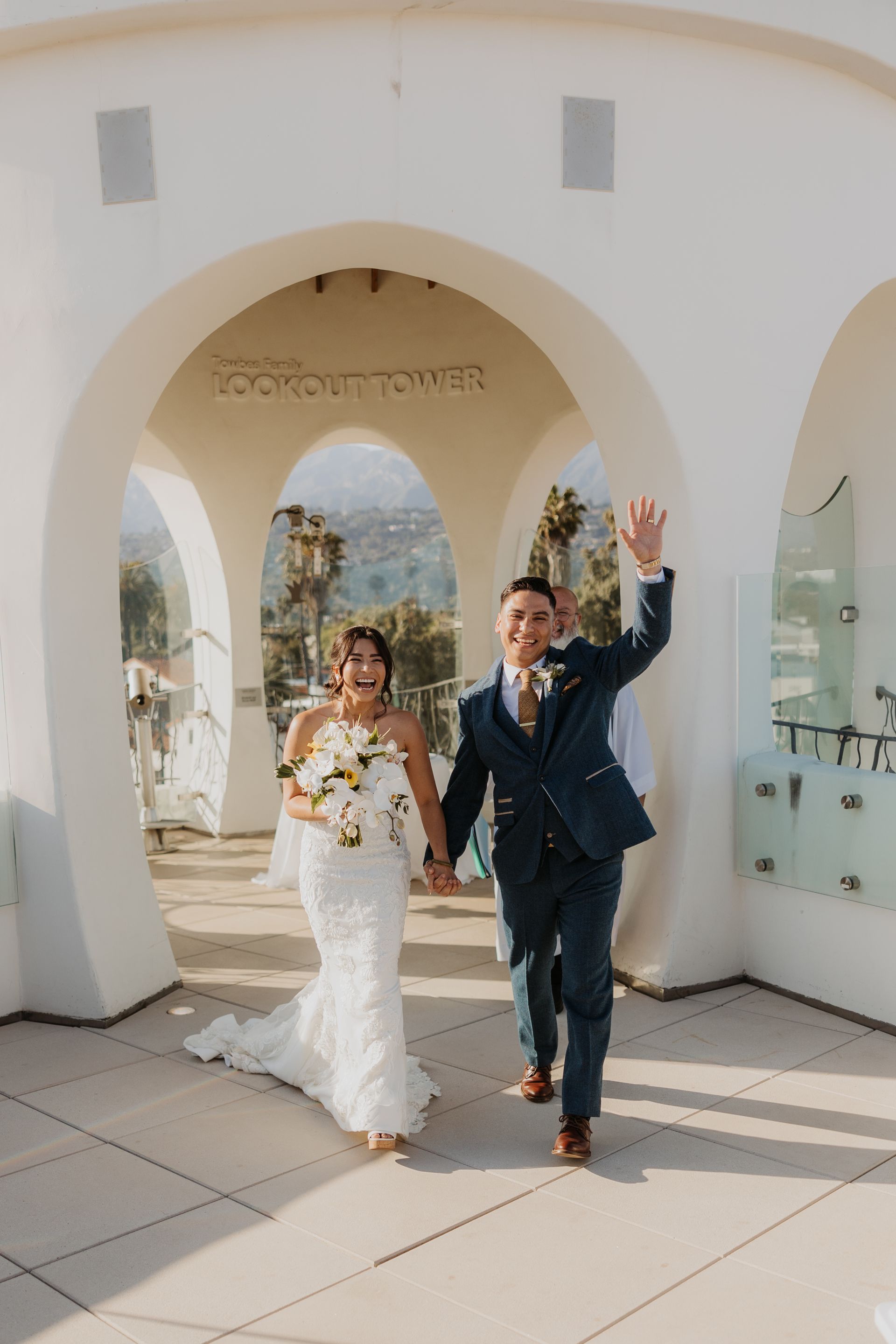 Bride and groom in wedding attire walking under a white archway, smiling and waving outdoors