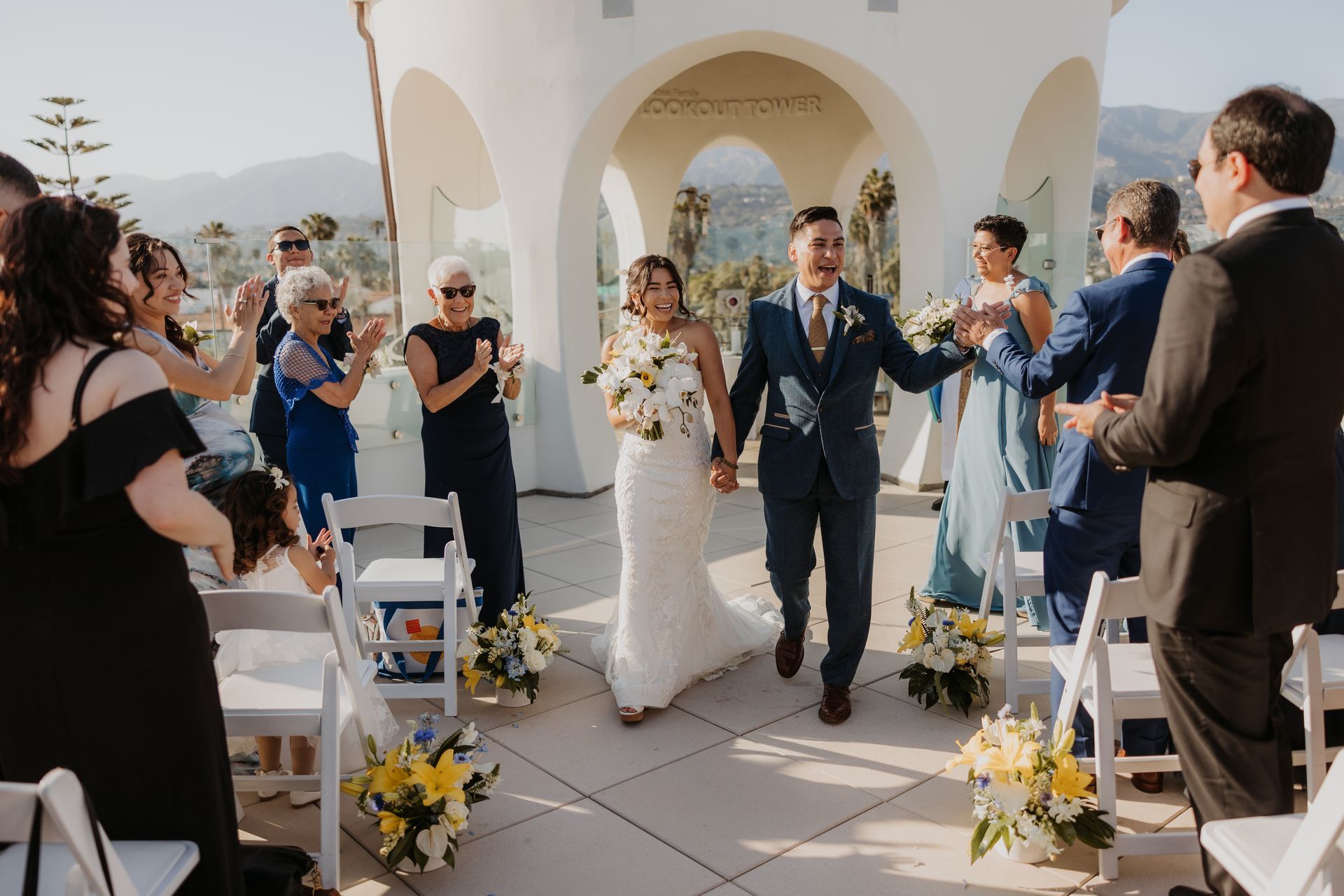 Bride and groom walk down an outdoor aisle at a sunlit wedding ceremony with guests applauding