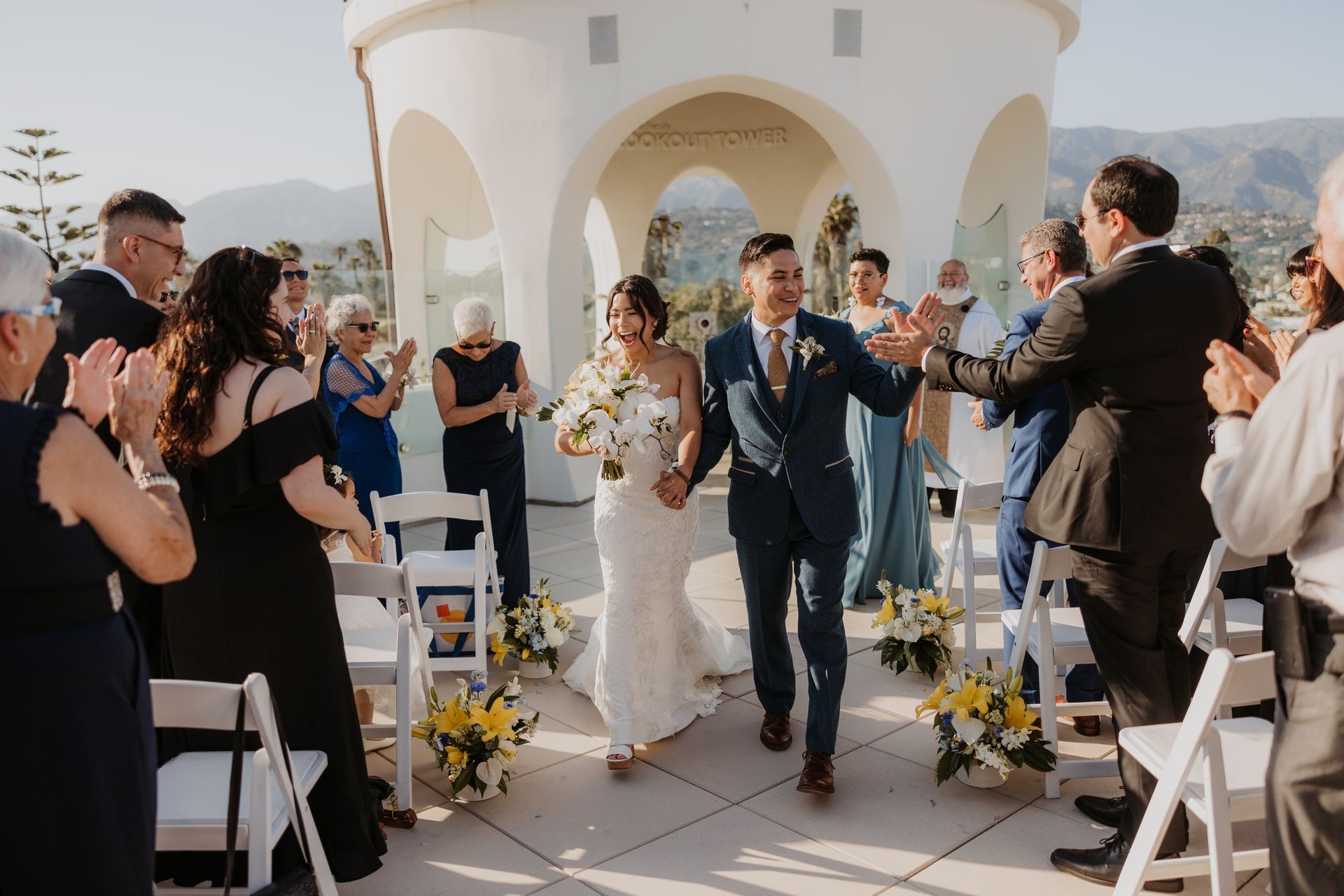 Outdoor wedding ceremony with bride and groom walking between applauding guests under a white arch