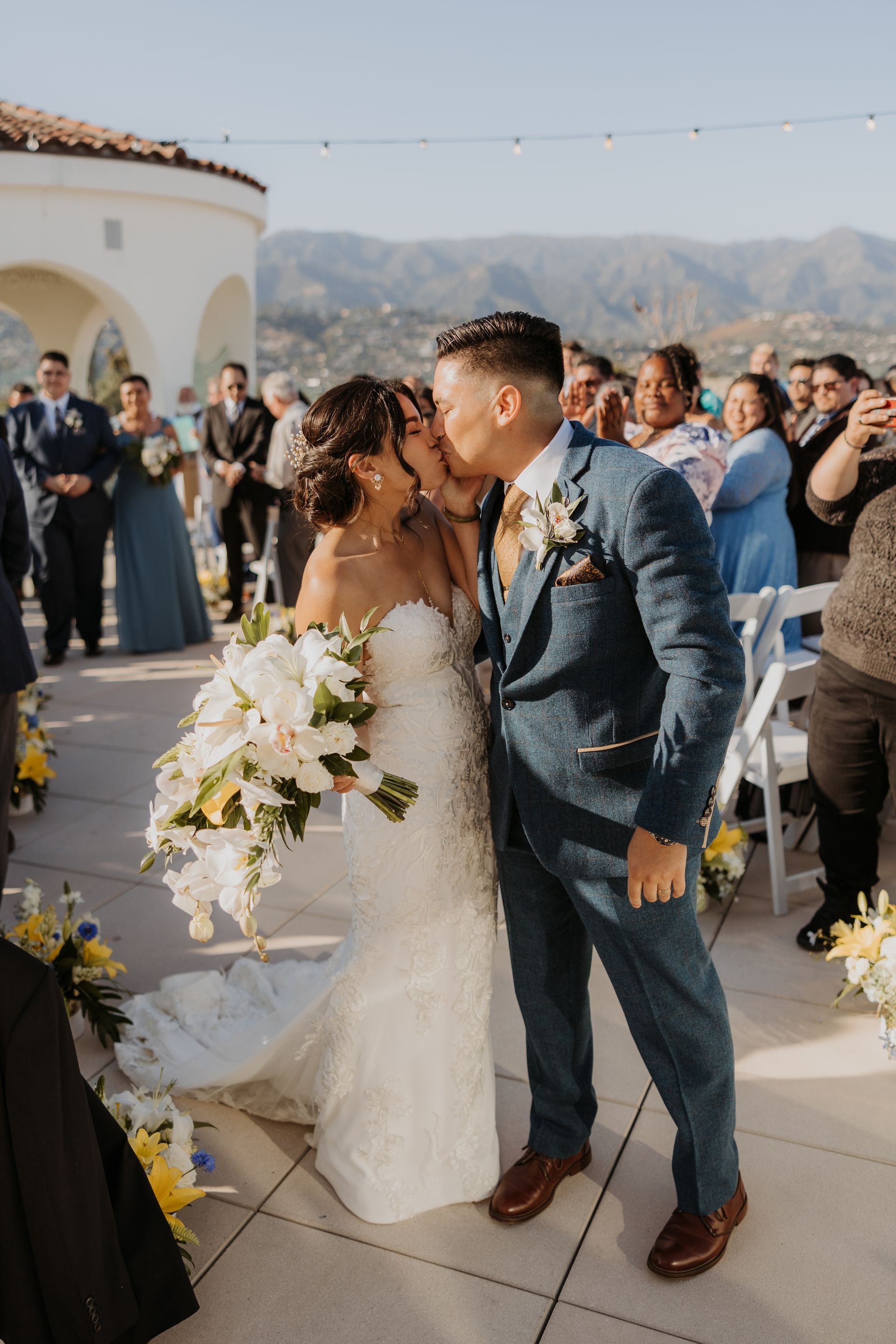 Bride and groom kissing at an outdoor wedding with guests applauding and mountains in the background
