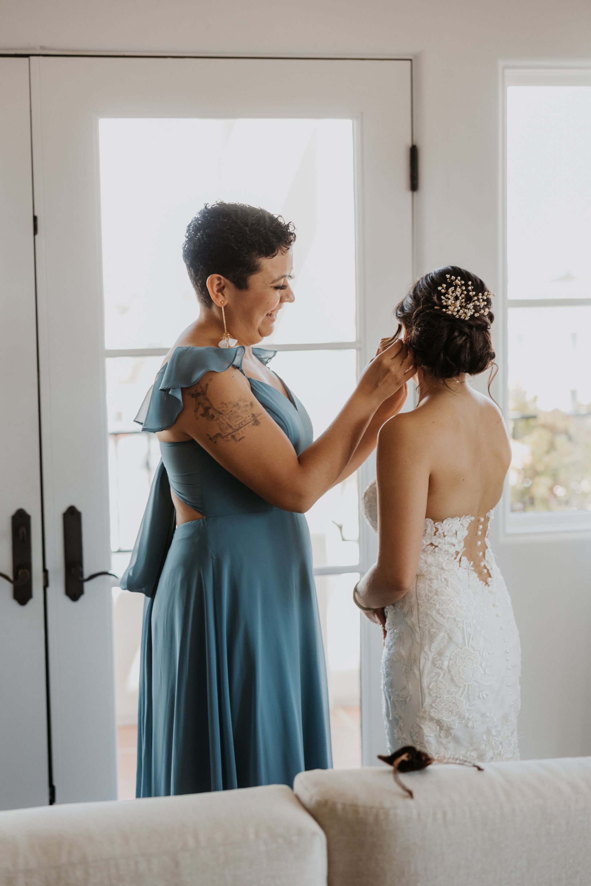 Two women in formal dresses adjusting each other’s attire by a bright doorway.