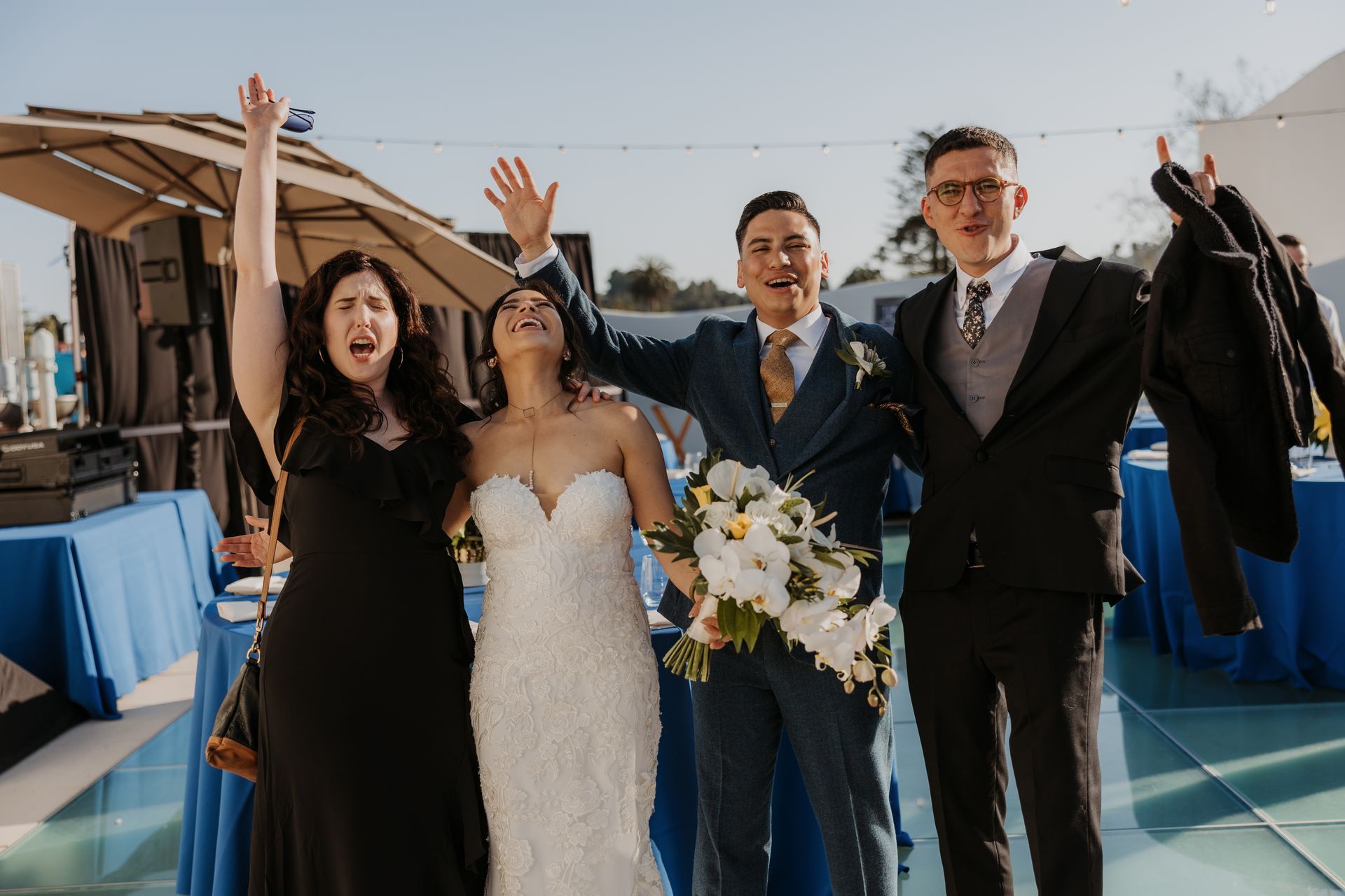 Wedding couple and two guests celebrating outdoors with raised arms and a bouquet