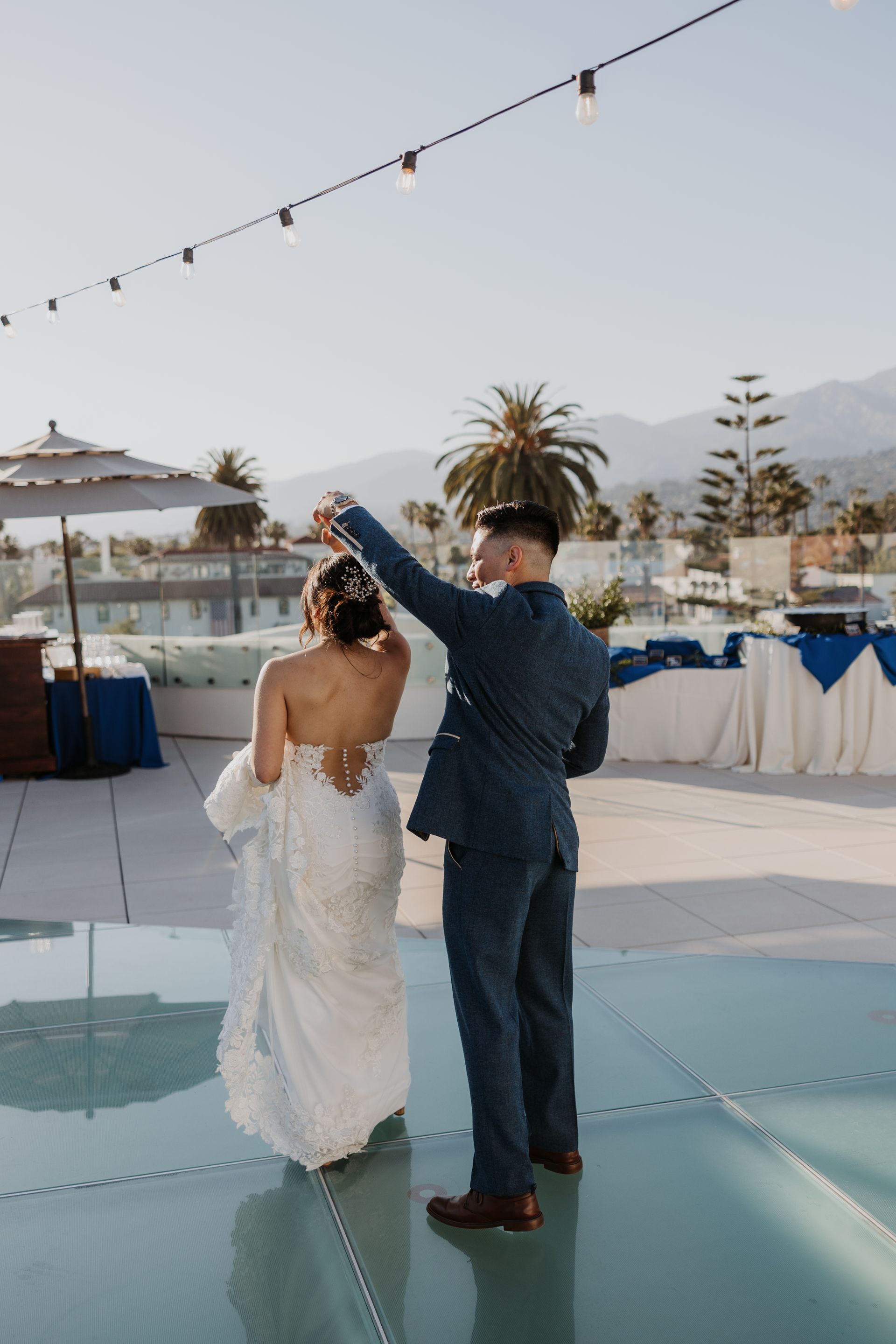Couple dancing on a glass platform at an outdoor wedding reception with string lights and palm trees