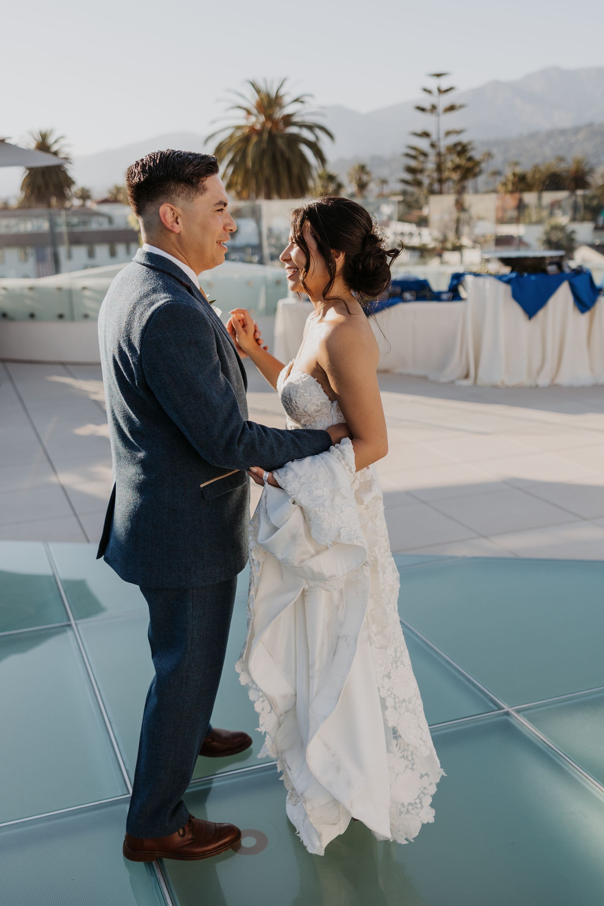 Couple in formal attire holding hands on a reflective rooftop terrace at an outdoor wedding reception