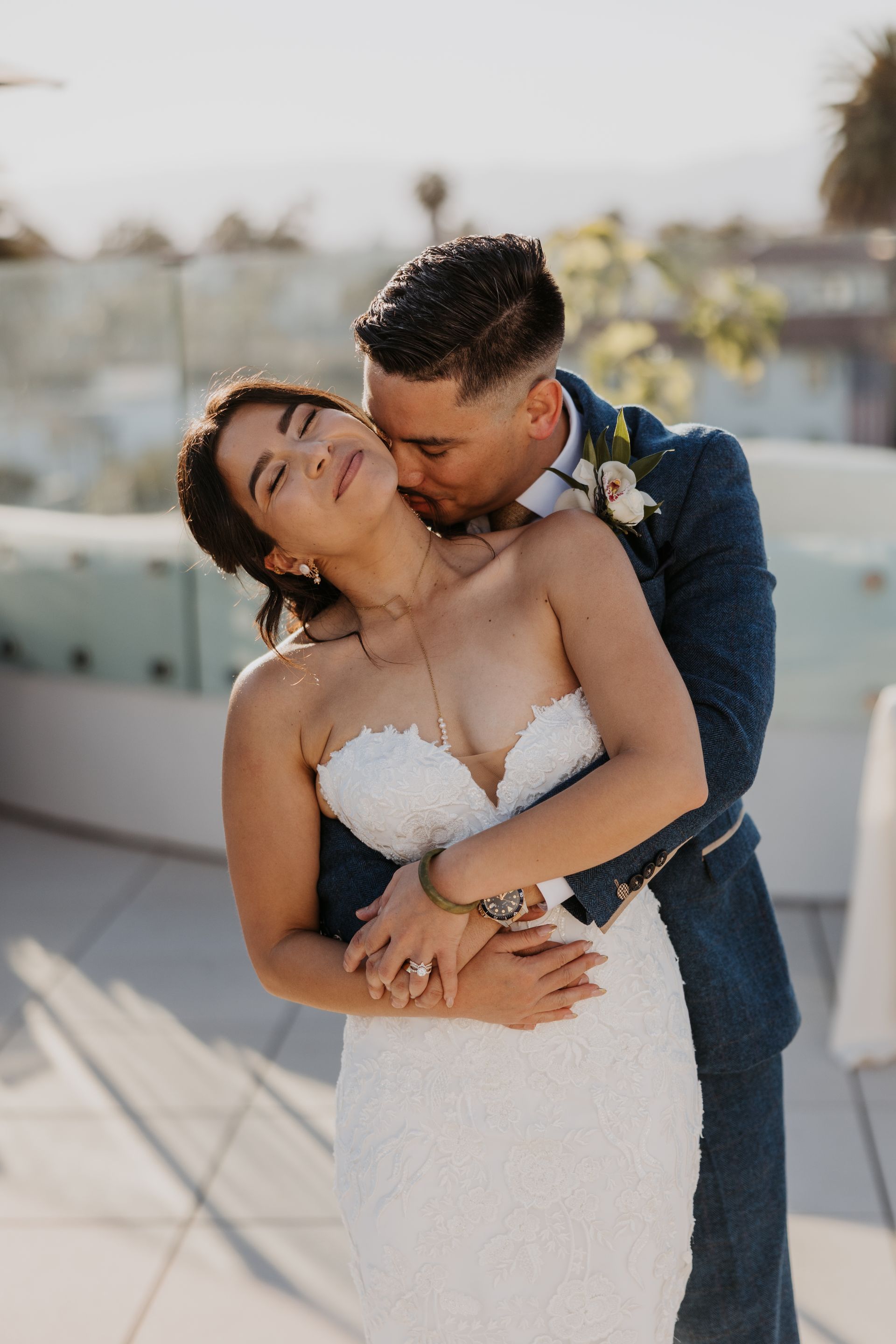 Couple embracing outdoors, groom kissing bride’s cheek as she smiles in a white dress.