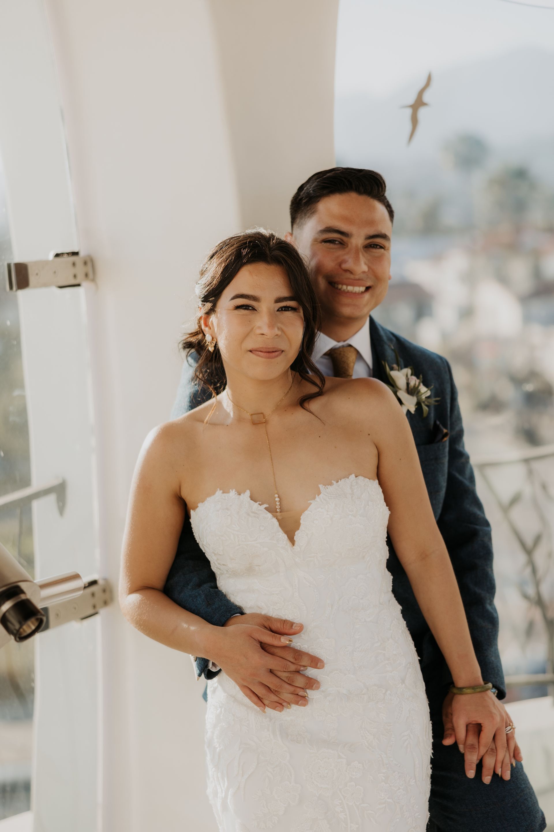 Bride in a white dress and groom in a dark suit standing together indoors, smiling and holding hands.