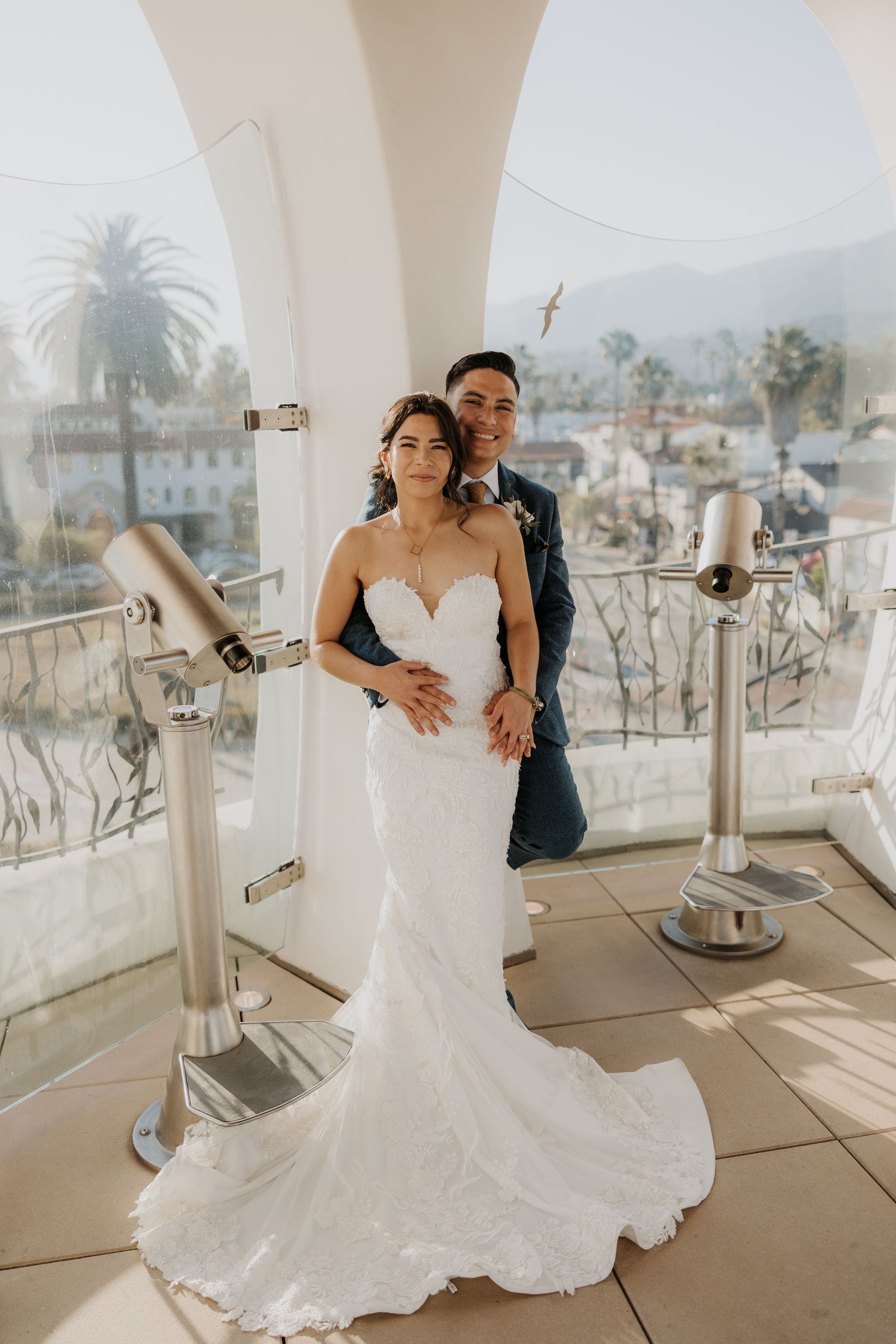 Bride and groom posing on a balcony in wedding attire with city view behind them