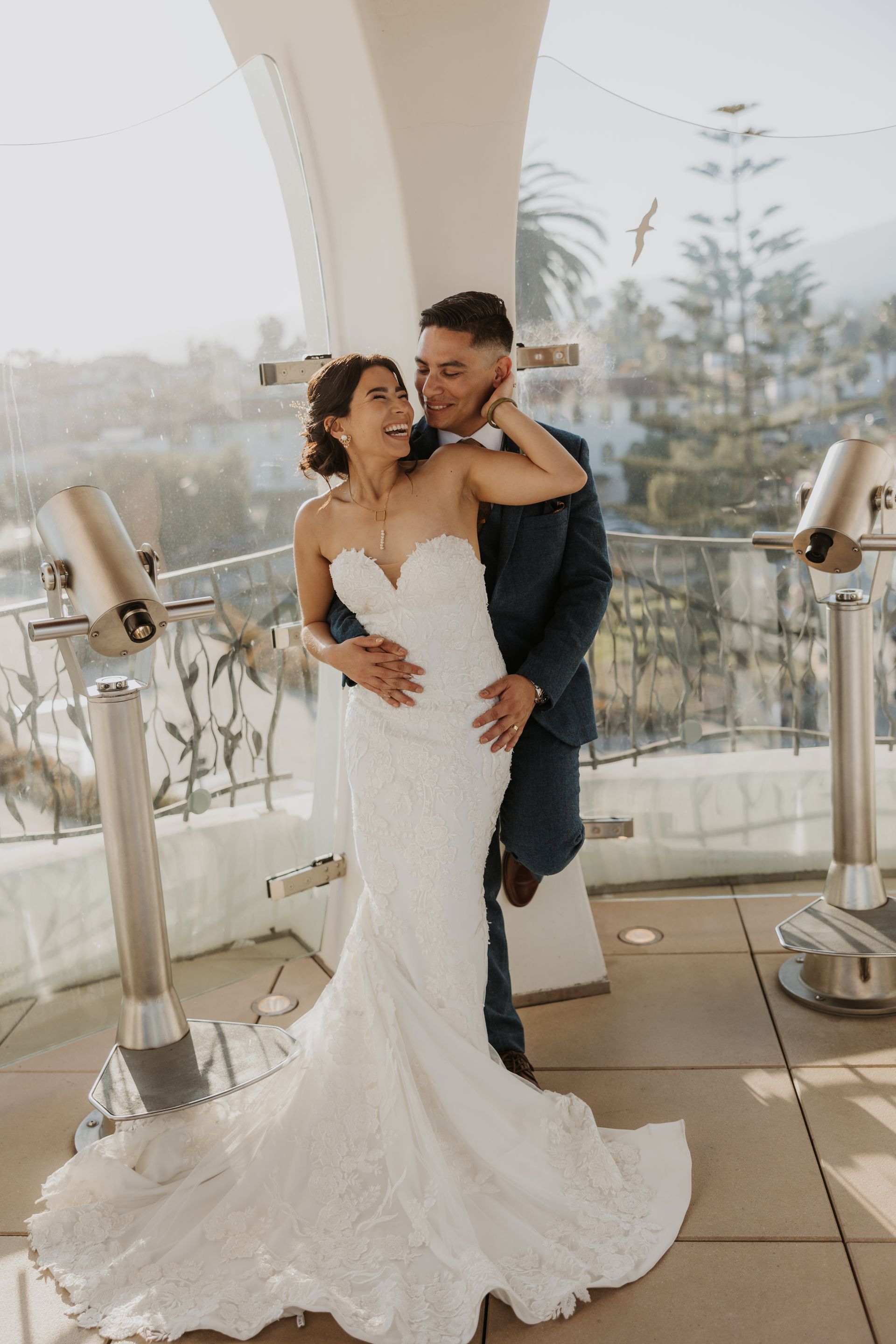 Bride in white gown being held from behind by groom on a balcony, with city view and soft daylight.