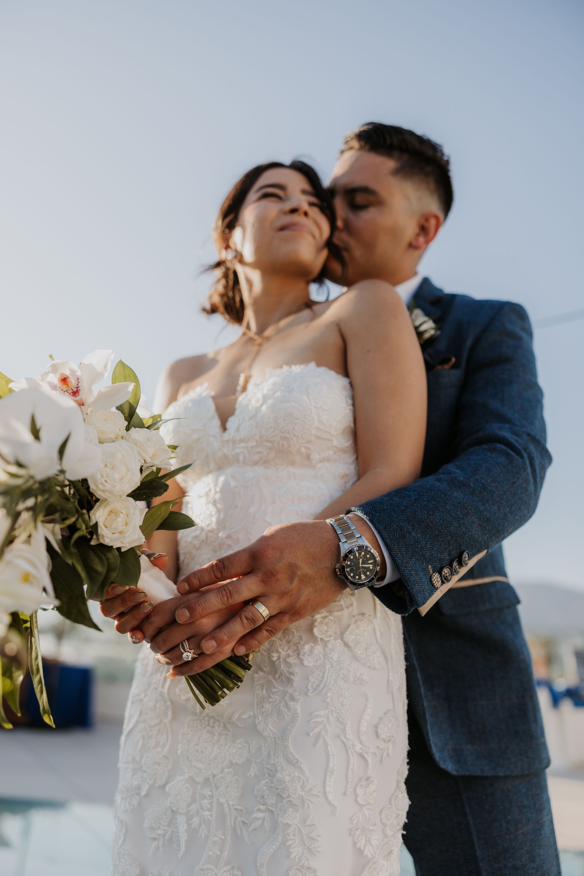 Bride and groom embracing outdoors, holding a white bouquet under a clear sky