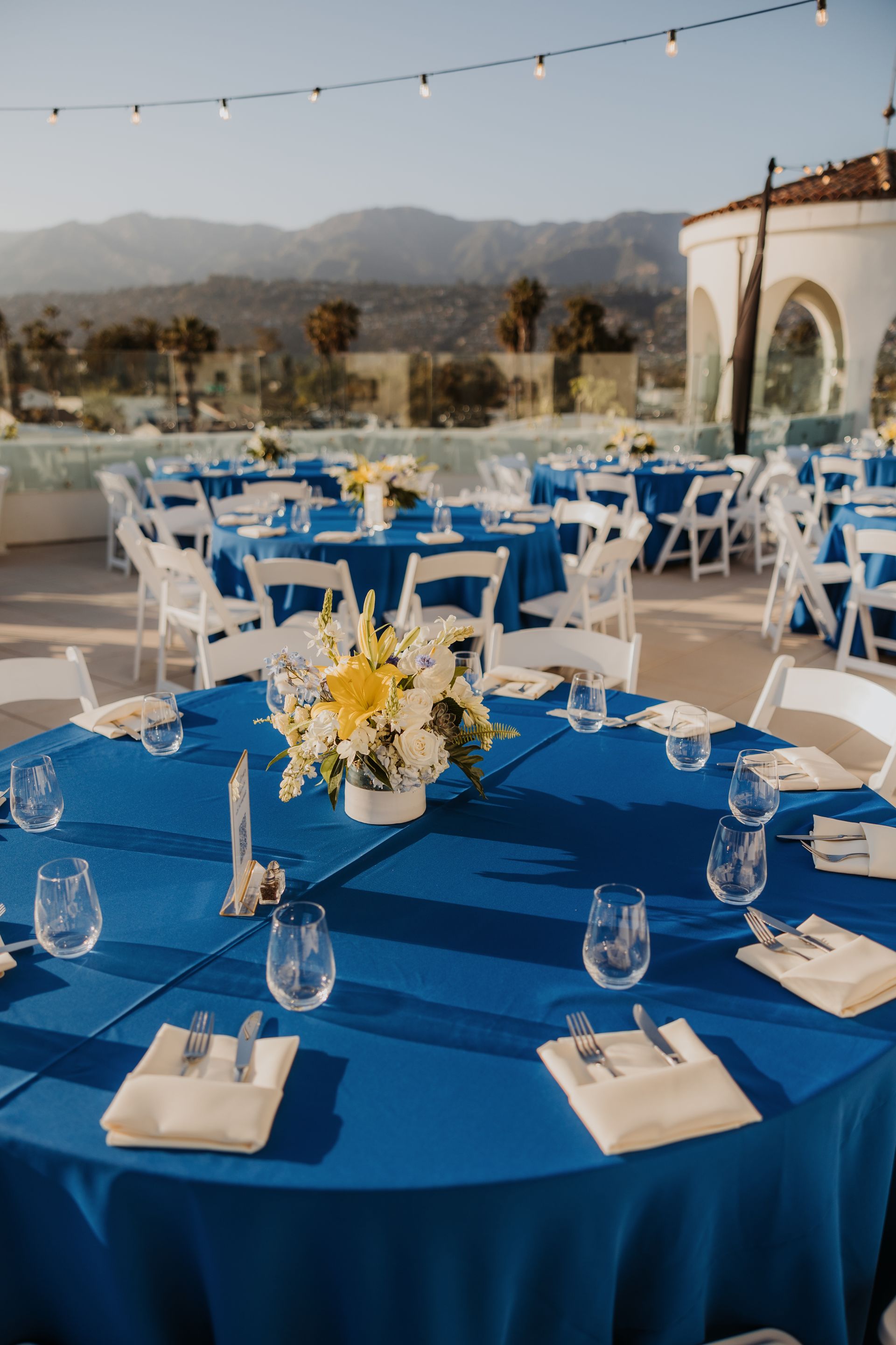 Outdoor event tables with blue tablecloths and white chairs, set against mountains at sunset