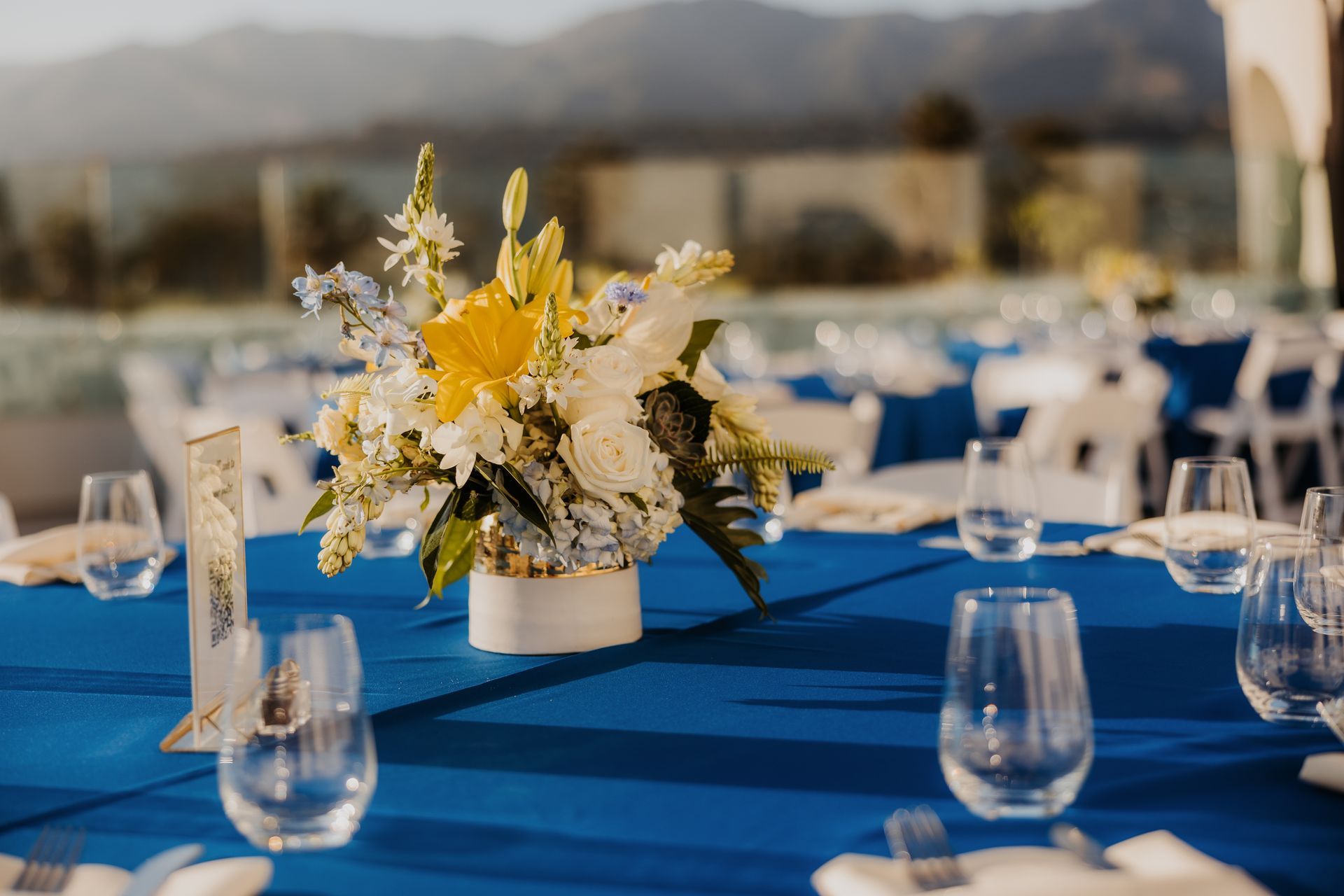 Blue table setting with white-and-yellow floral centerpiece and glassware at an outdoor event