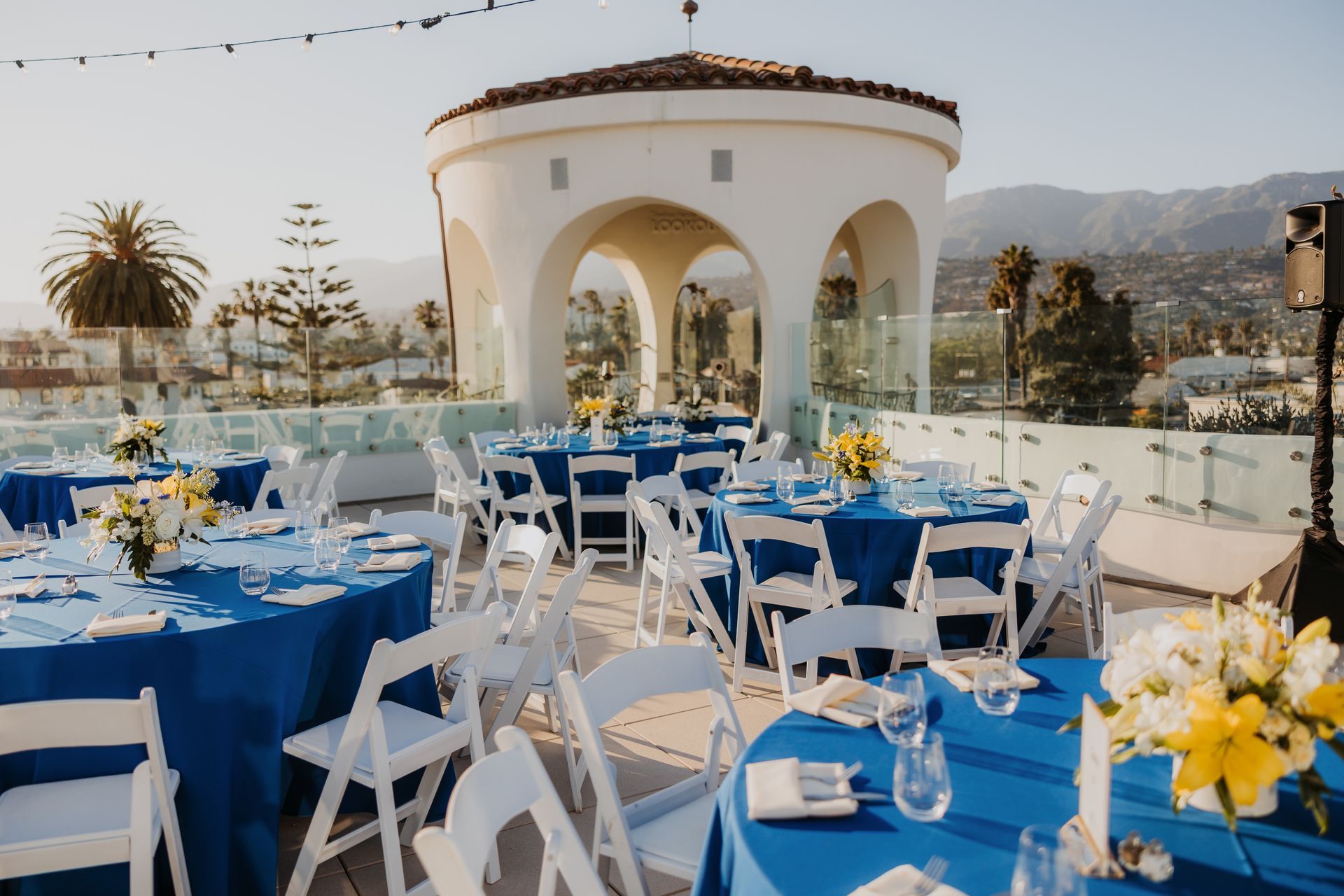 Outdoor event tables with blue tablecloths and white chairs beside a white pavilion with mountains in the background