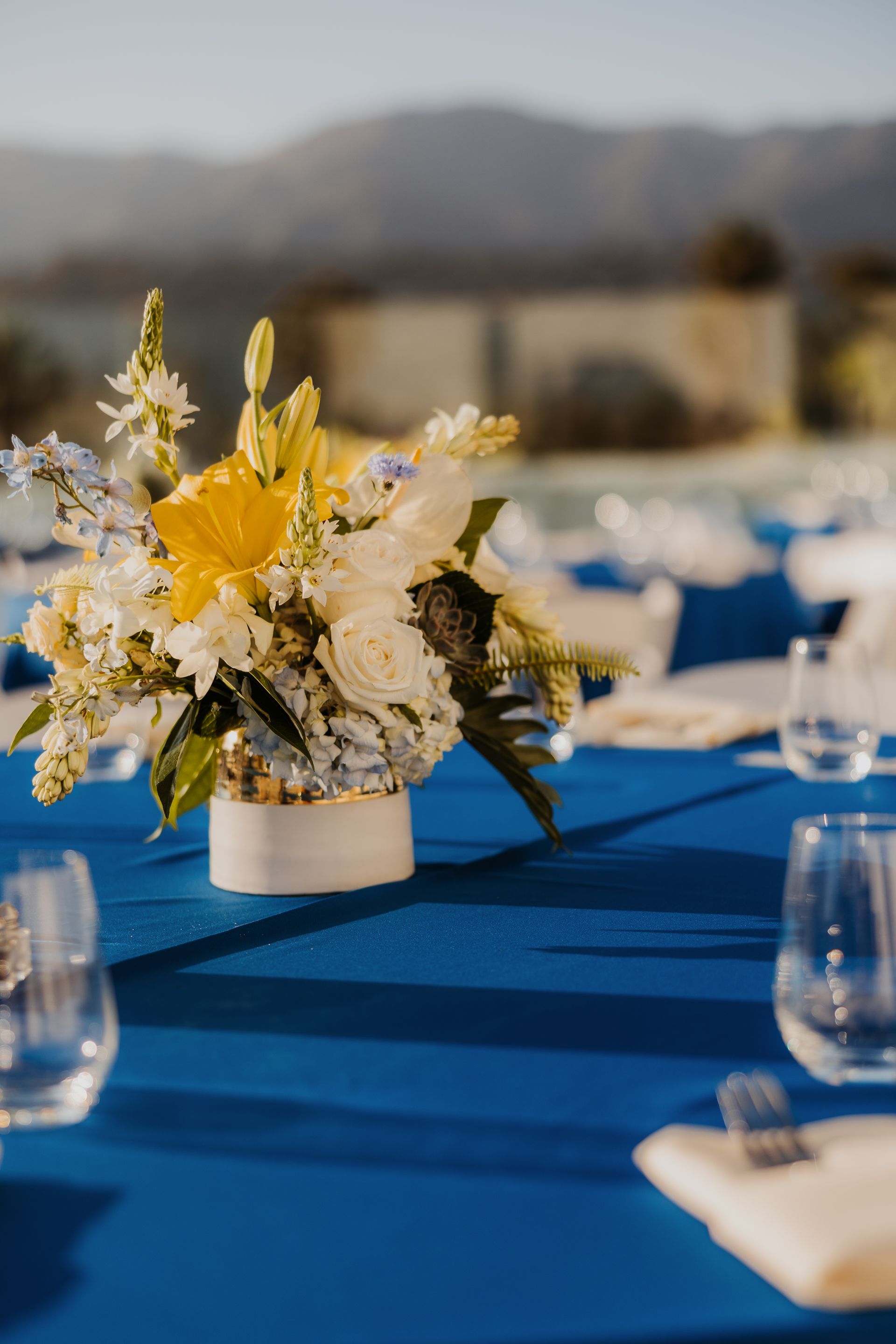 White floral centerpiece with yellow blooms on a blue outdoor dining table at sunset