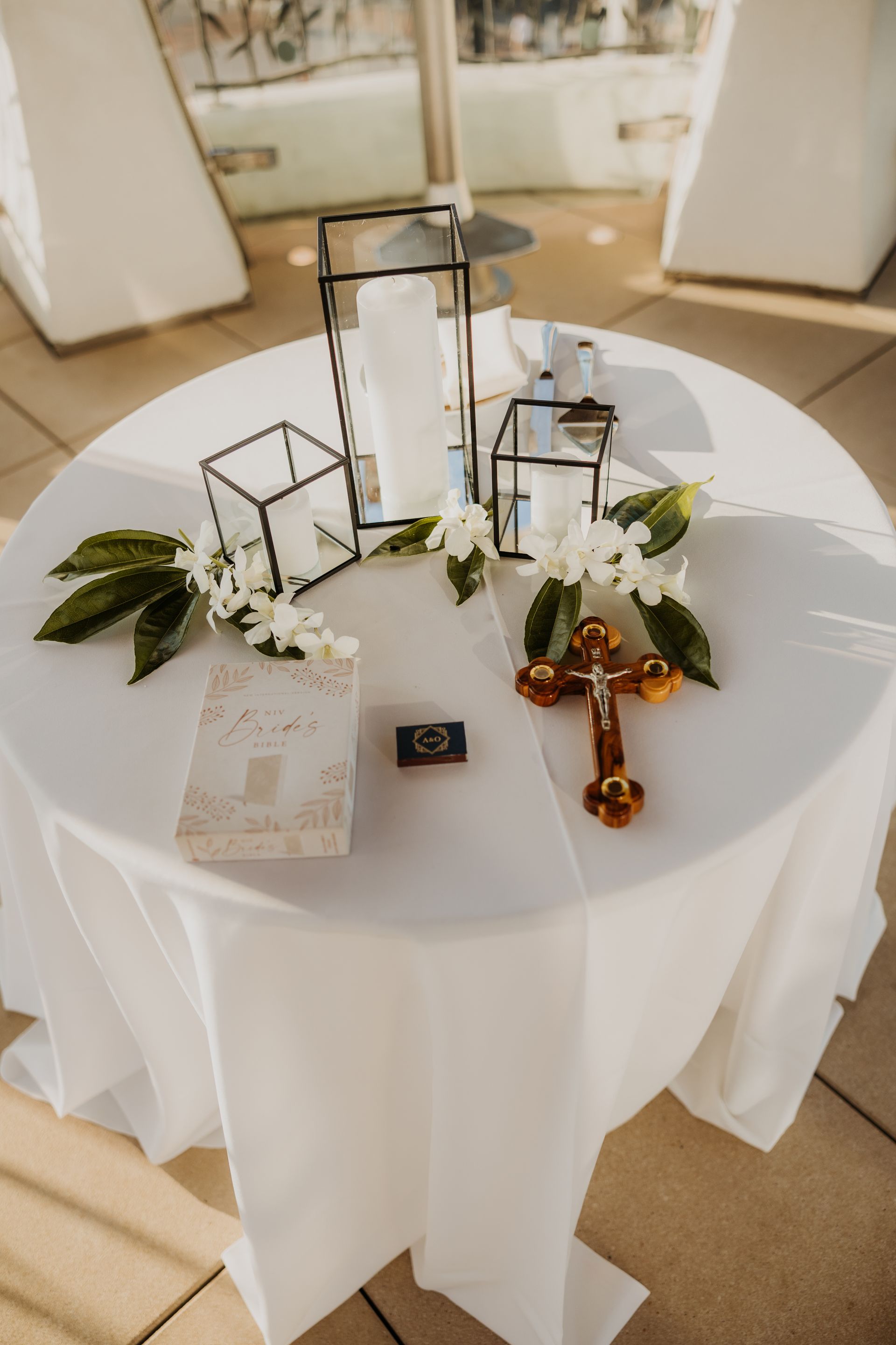 White round table with a glass lantern centerpiece, flowers, a booklet, and a cross at a wedding ceremony