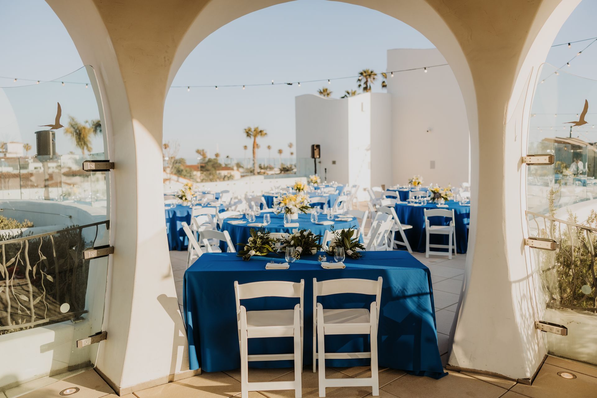 Blue-draped outdoor dining tables beneath white arches overlooking the sea