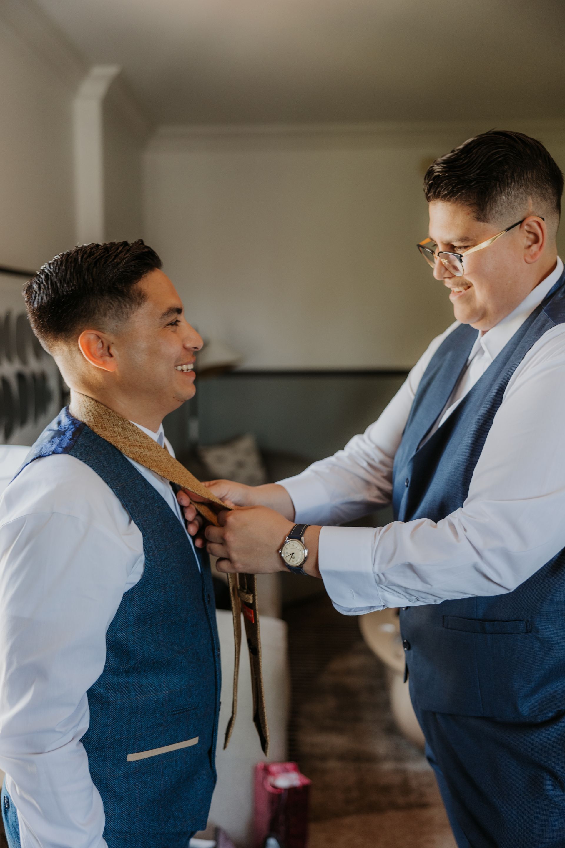 Two men in blue vests adjusting a tie in a softly lit room