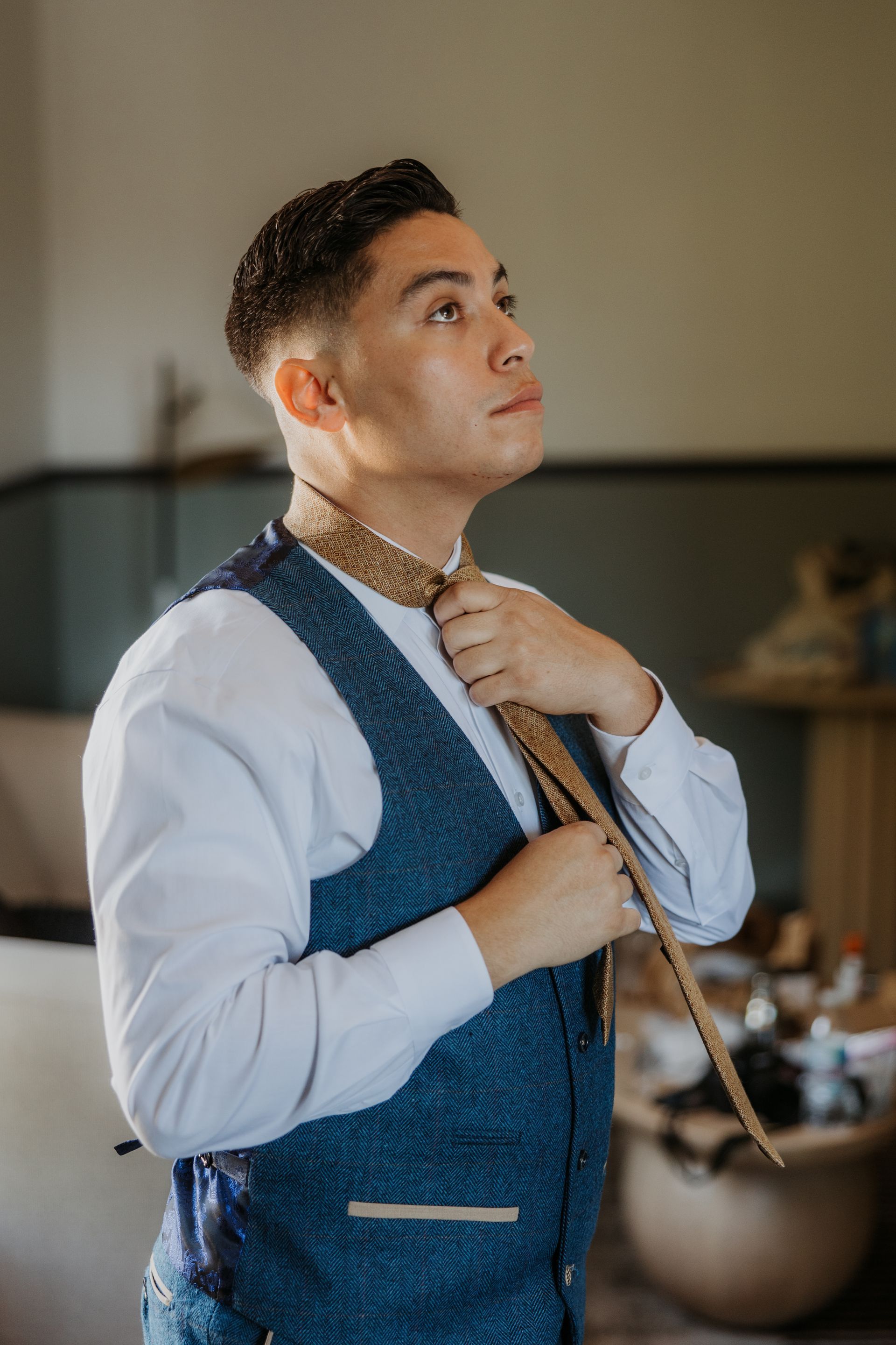 Person adjusting a tie in a blue vest and white shirt, standing indoors before a mirror