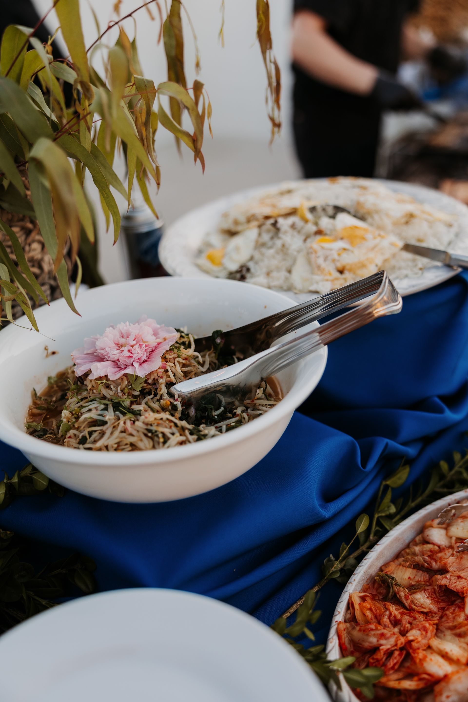 Buffet table with bowls of salad, rice, and kimchi on blue cloth, with flowers and serving tongs