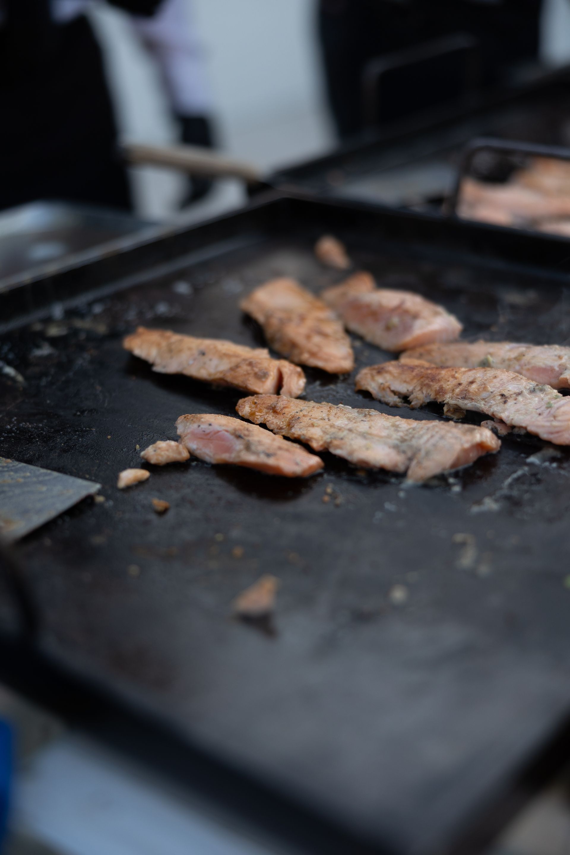 Slices of meat cooking on a black griddle with a spatula in the background