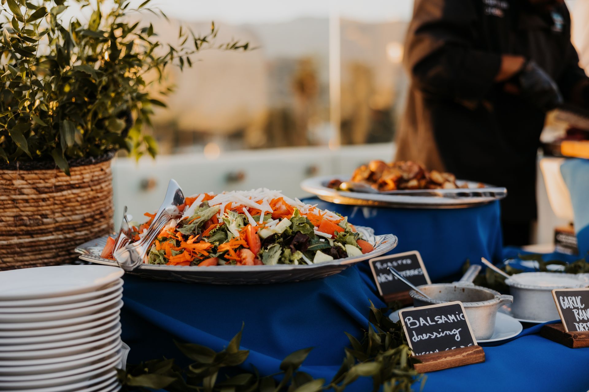 Buffet table with blue bowls of salad, stacked plates, and a chalkboard sign at an outdoor event