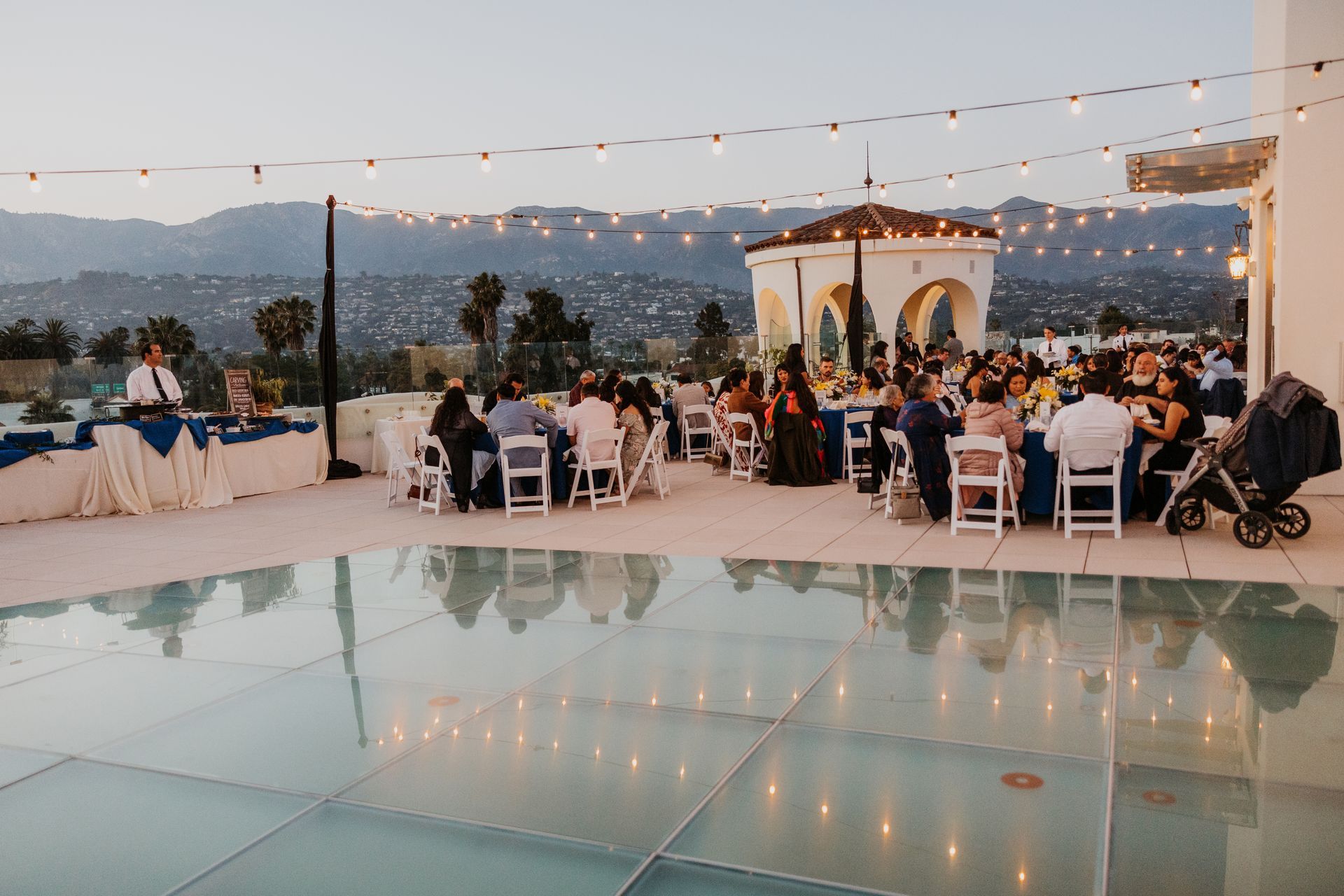 Outdoor evening wedding reception by a pool with seated guests, string lights, and a mountain backdrop