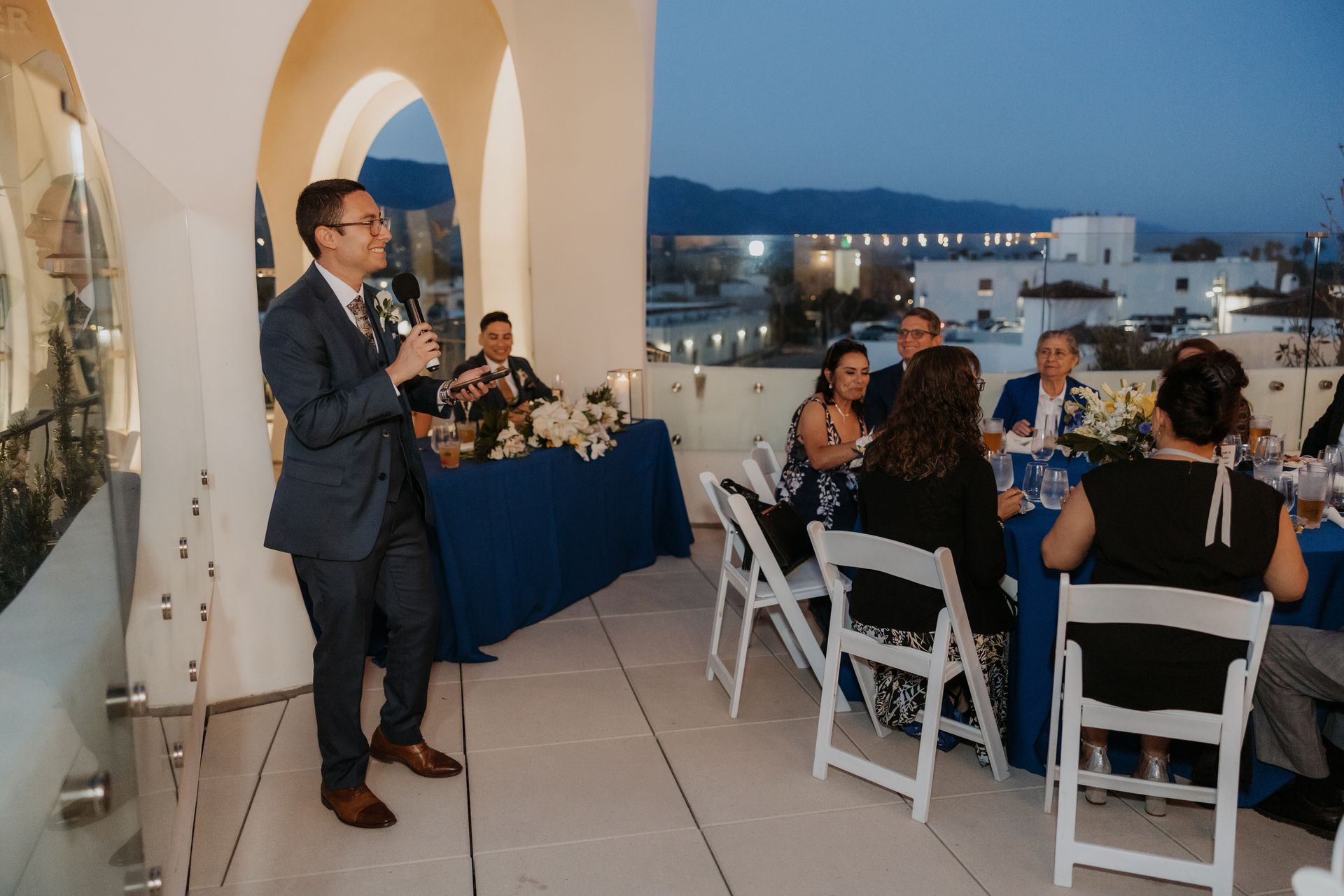 Man speaking at a rooftop evening reception beside seated guests and city lights