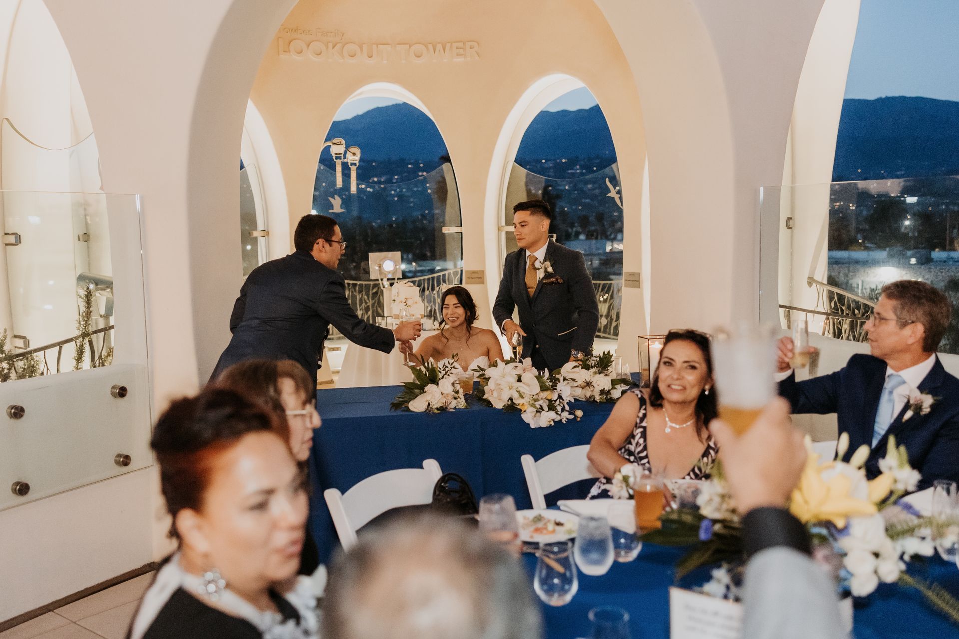 Dinner party on a terrace with guests seated at a blue-clothed table overlooking the sea